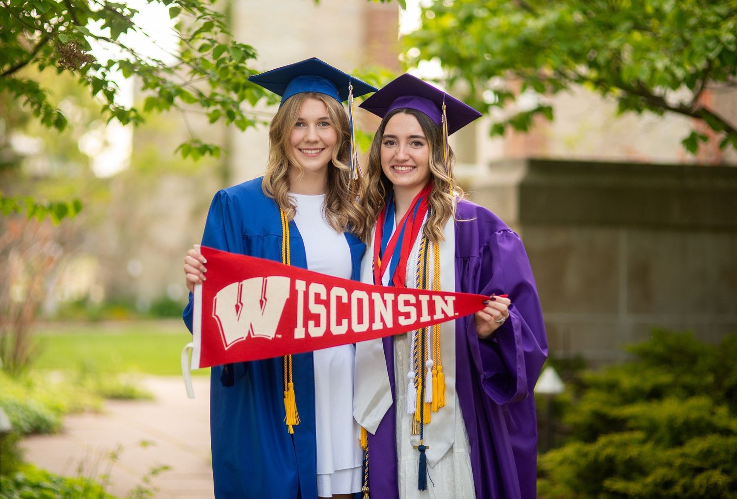 Two women in blue and purple graduation gowns and caps hold a red Wisconsin pennant outdoors among green trees.