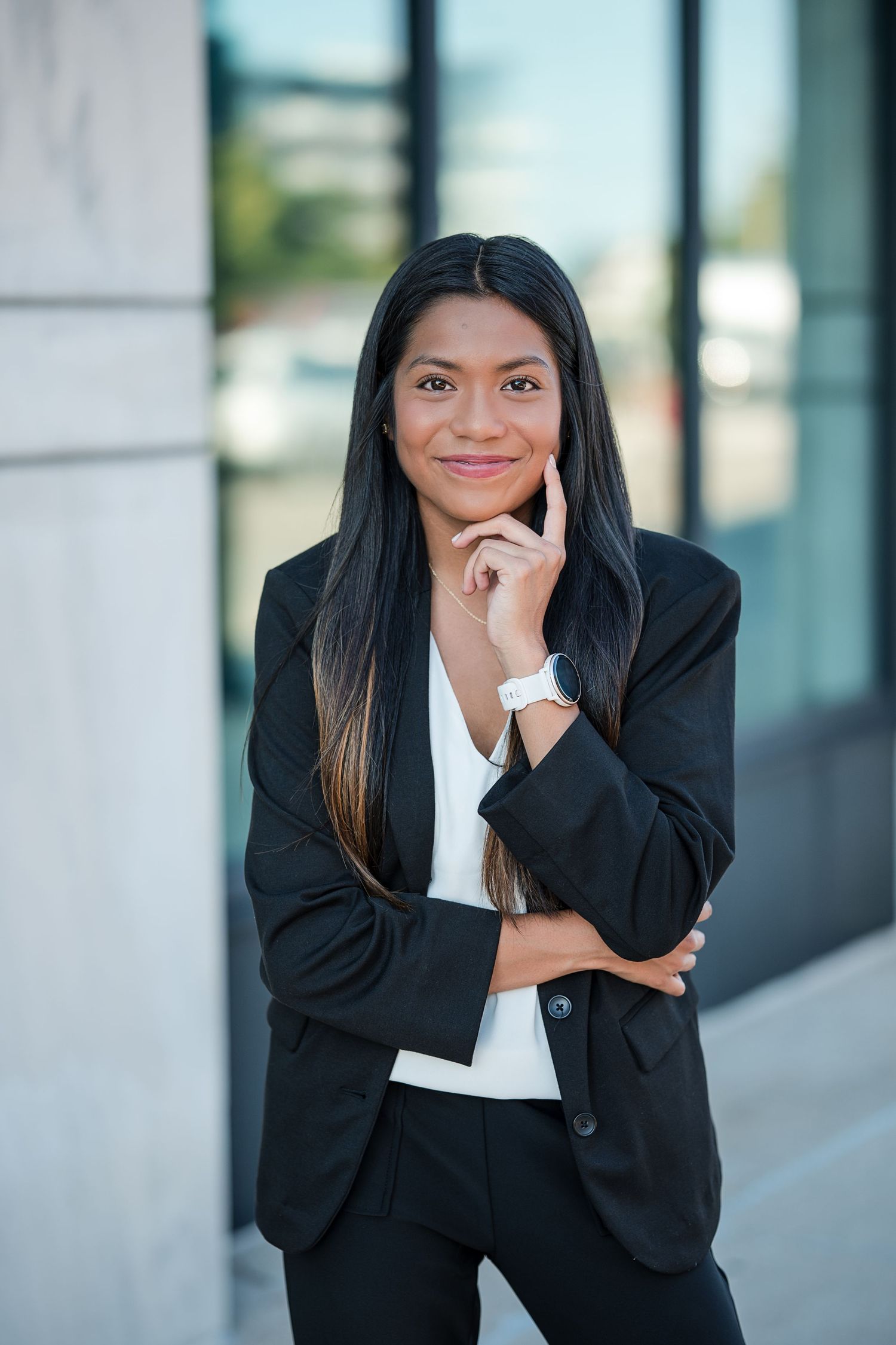A confident young businesswoman in a black suit poses thoughtfully with her hand on her chin outside an office building.
