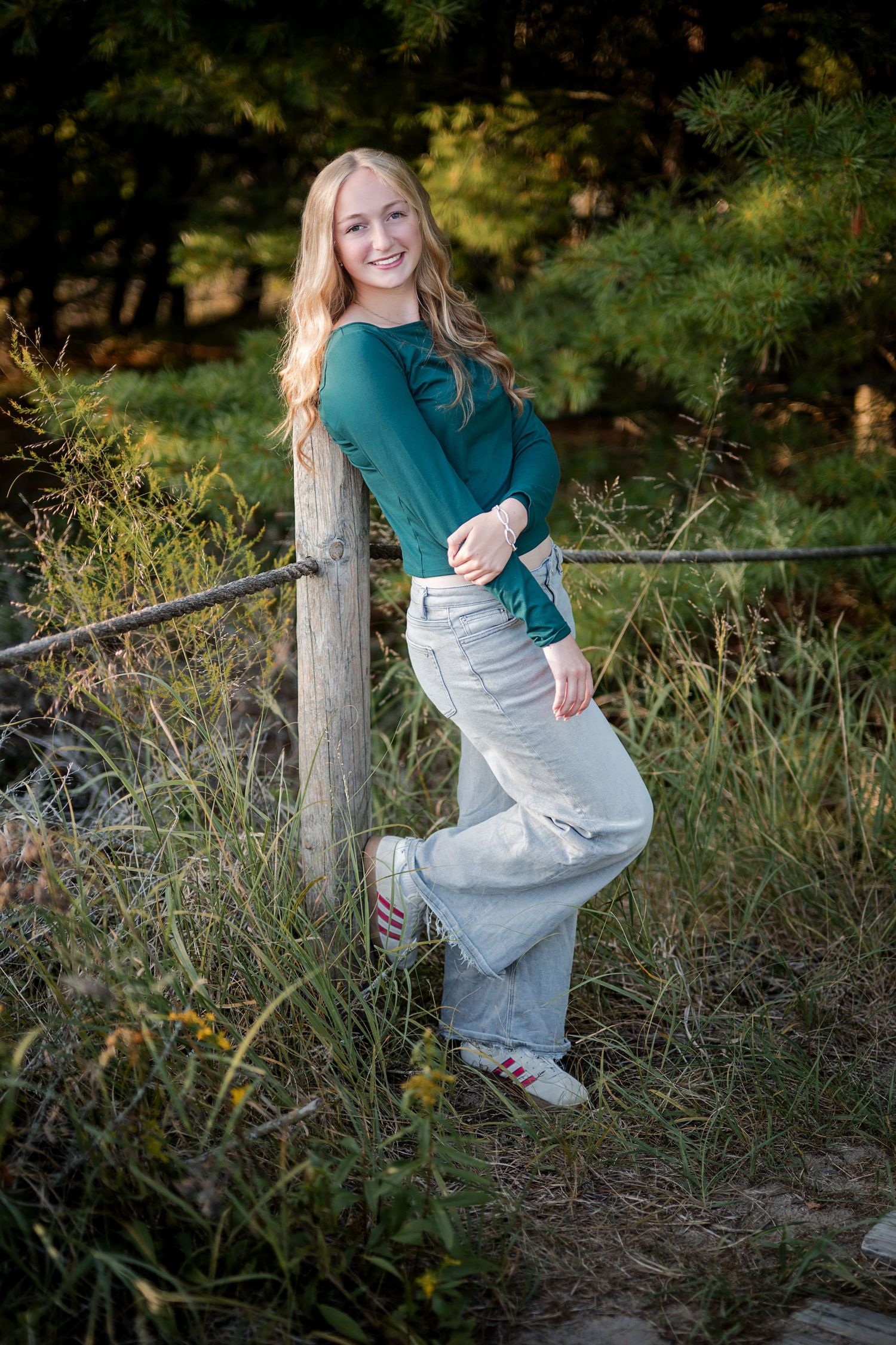 Smiling blonde teen girl in teal top and wide-leg pants leaning on wooden fence post in natural outdoor setting.