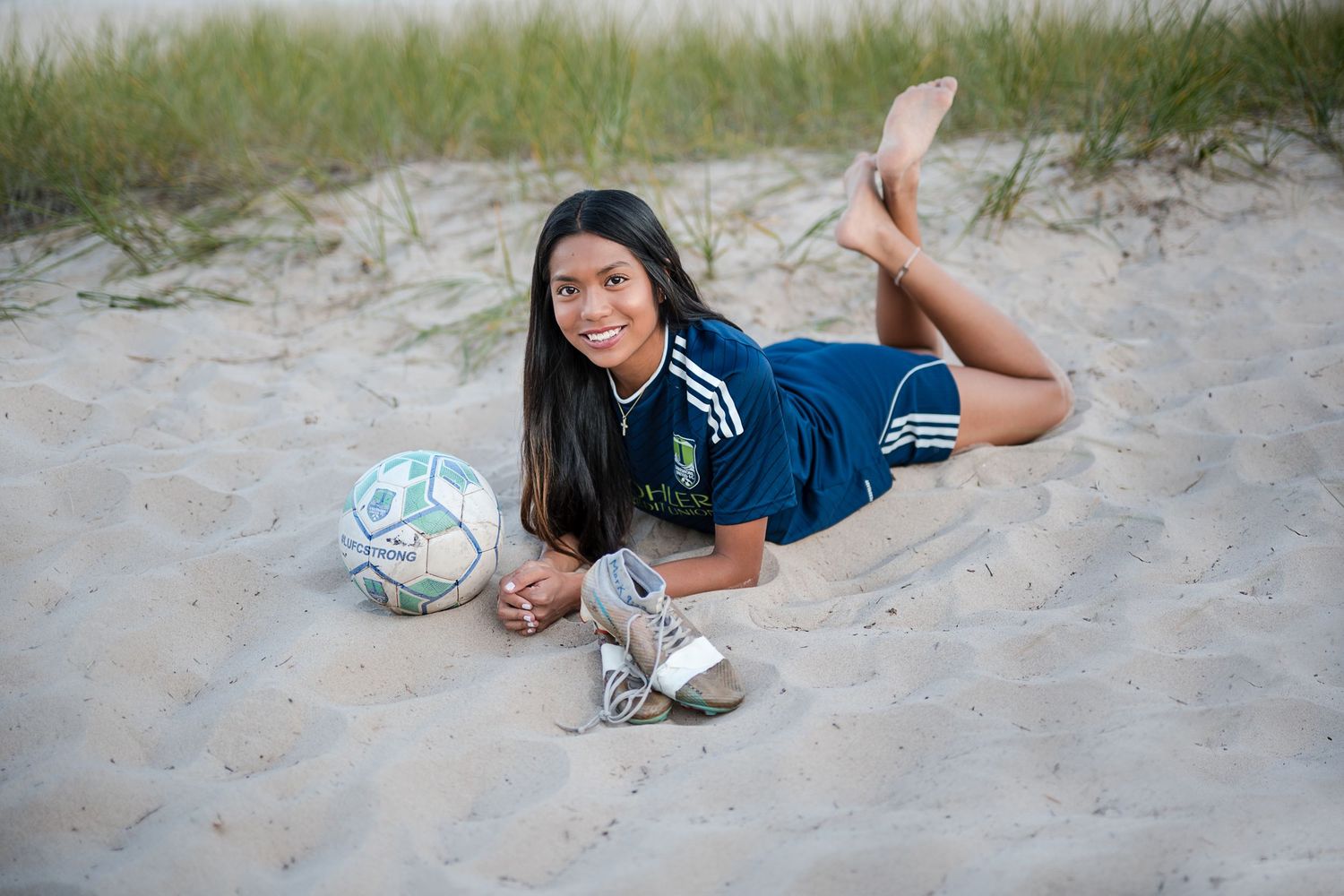 Young female soccer player lying on sandy beach in blue jersey, smiling with soccer ball and cleats nearby.