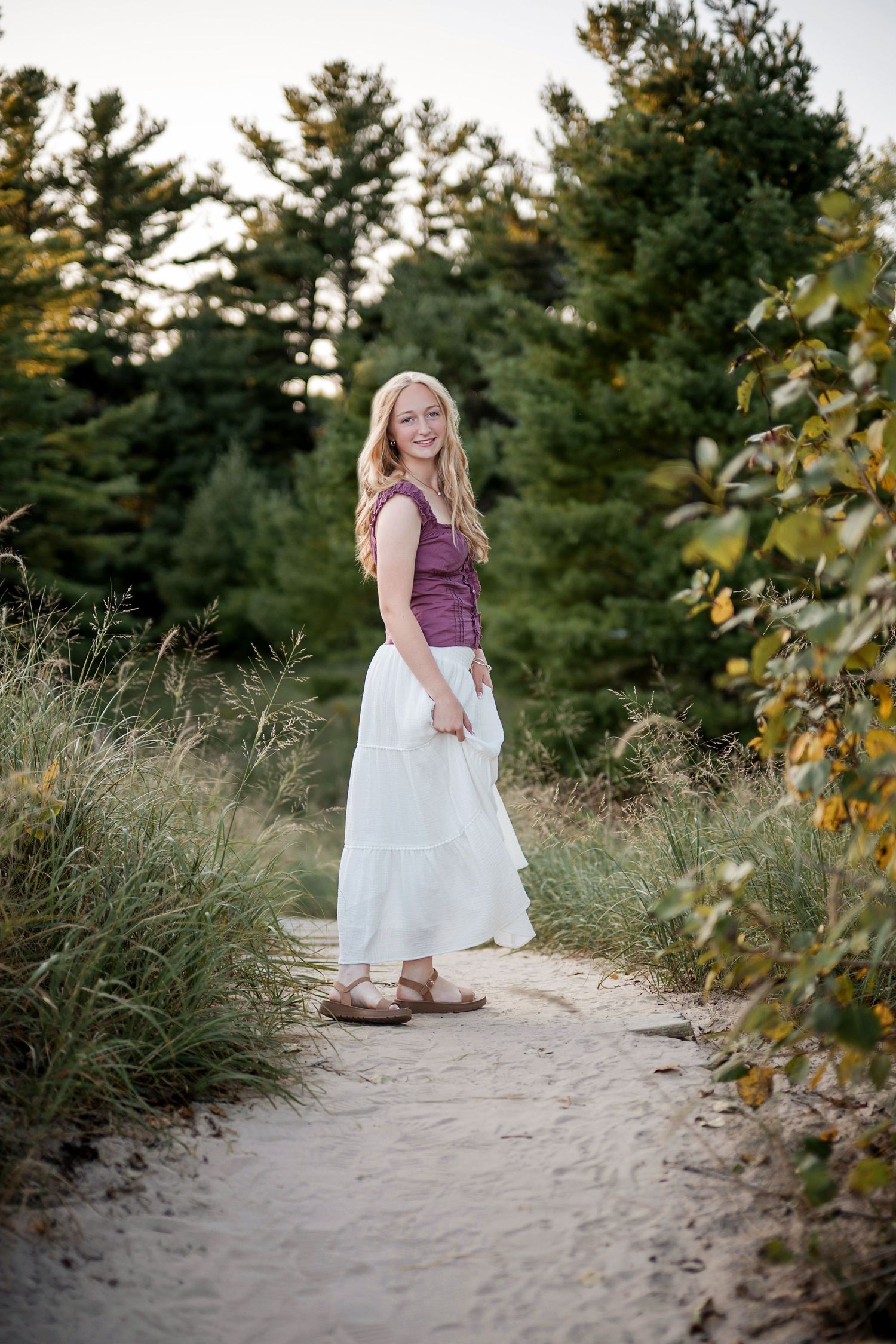 Smiling blonde woman in white skirt and purple top standing on a sandy path surrounded by green trees.