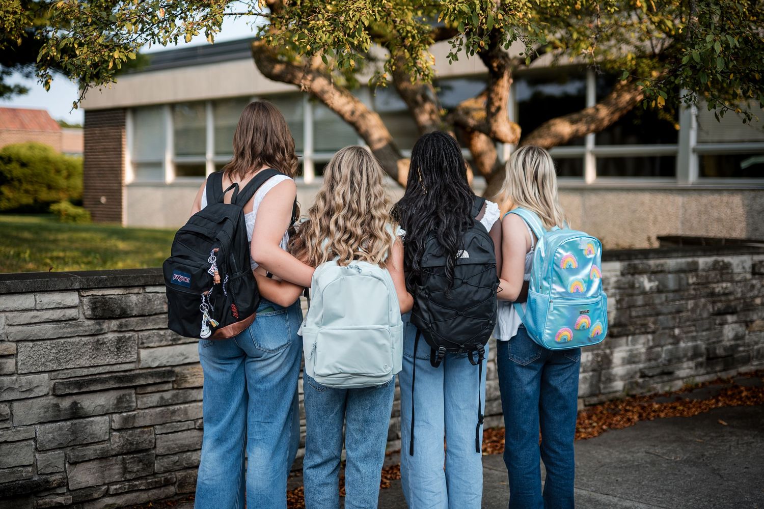 Four girls in jeans wearing backpacks stand with their backs to the camera beside a stone wall and a school building.