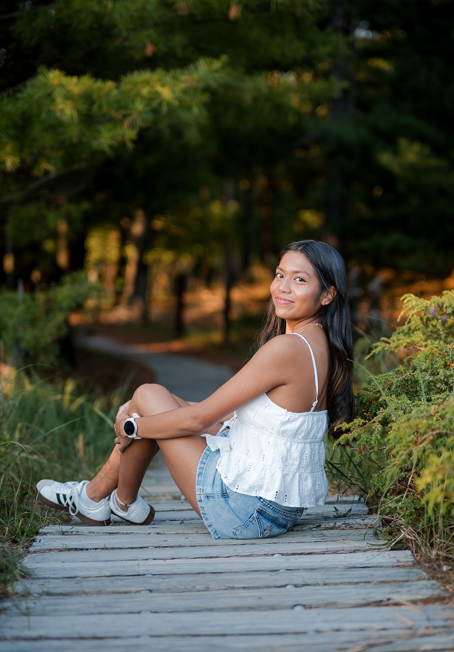 Young woman in white top and denim shorts sitting on a wooden boardwalk path surrounded by lush green trees.