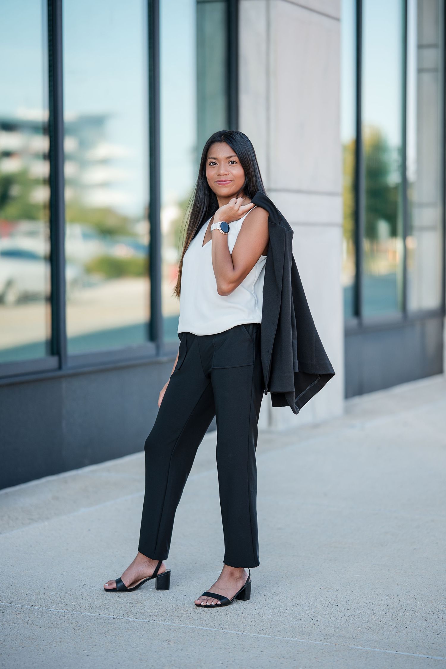 Young woman in black trousers, white top, and blazer over shoulder poses outdoors near modern building.
