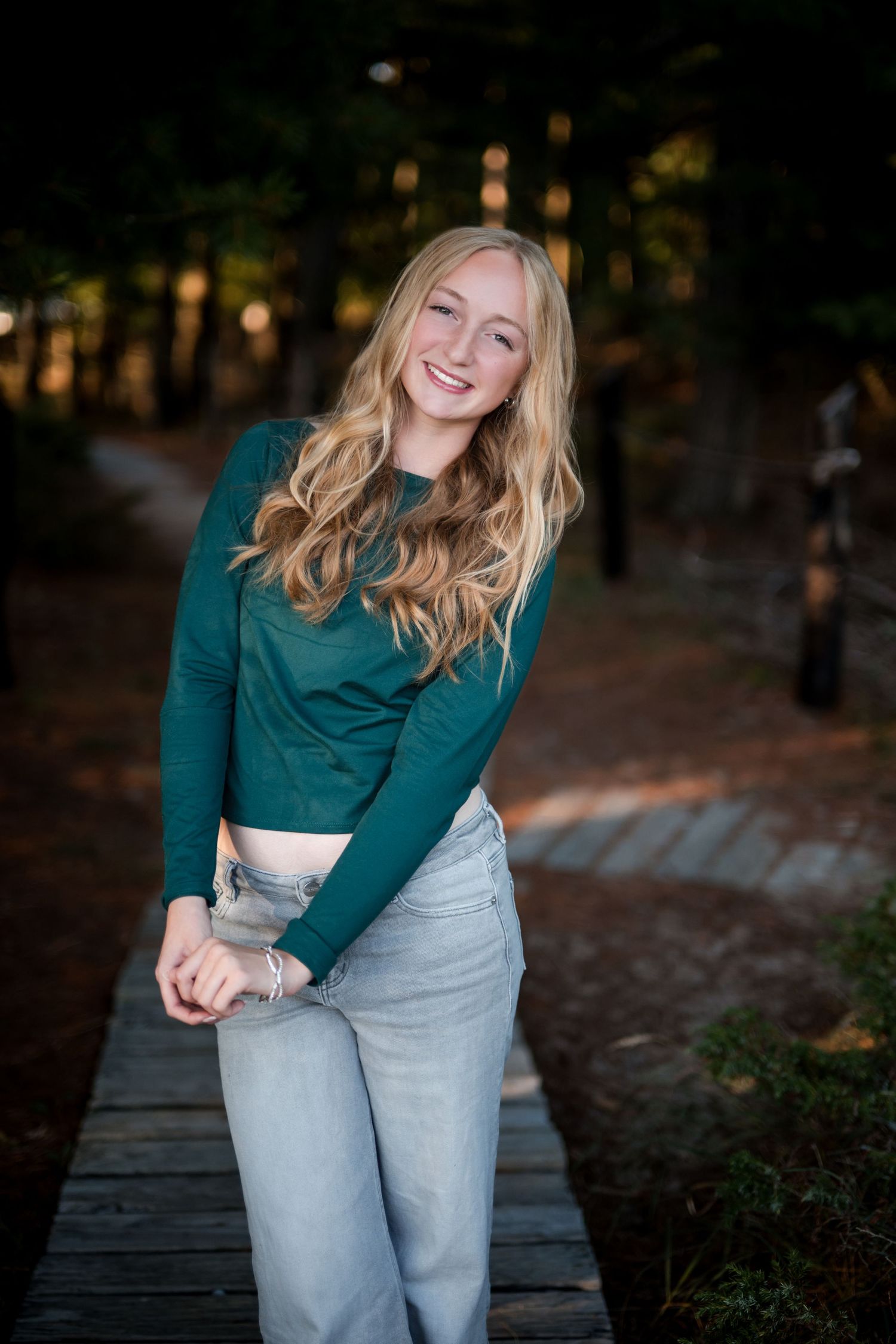Smiling teenage girl with blonde wavy hair wearing a green crop top and light jeans on a wooden path.