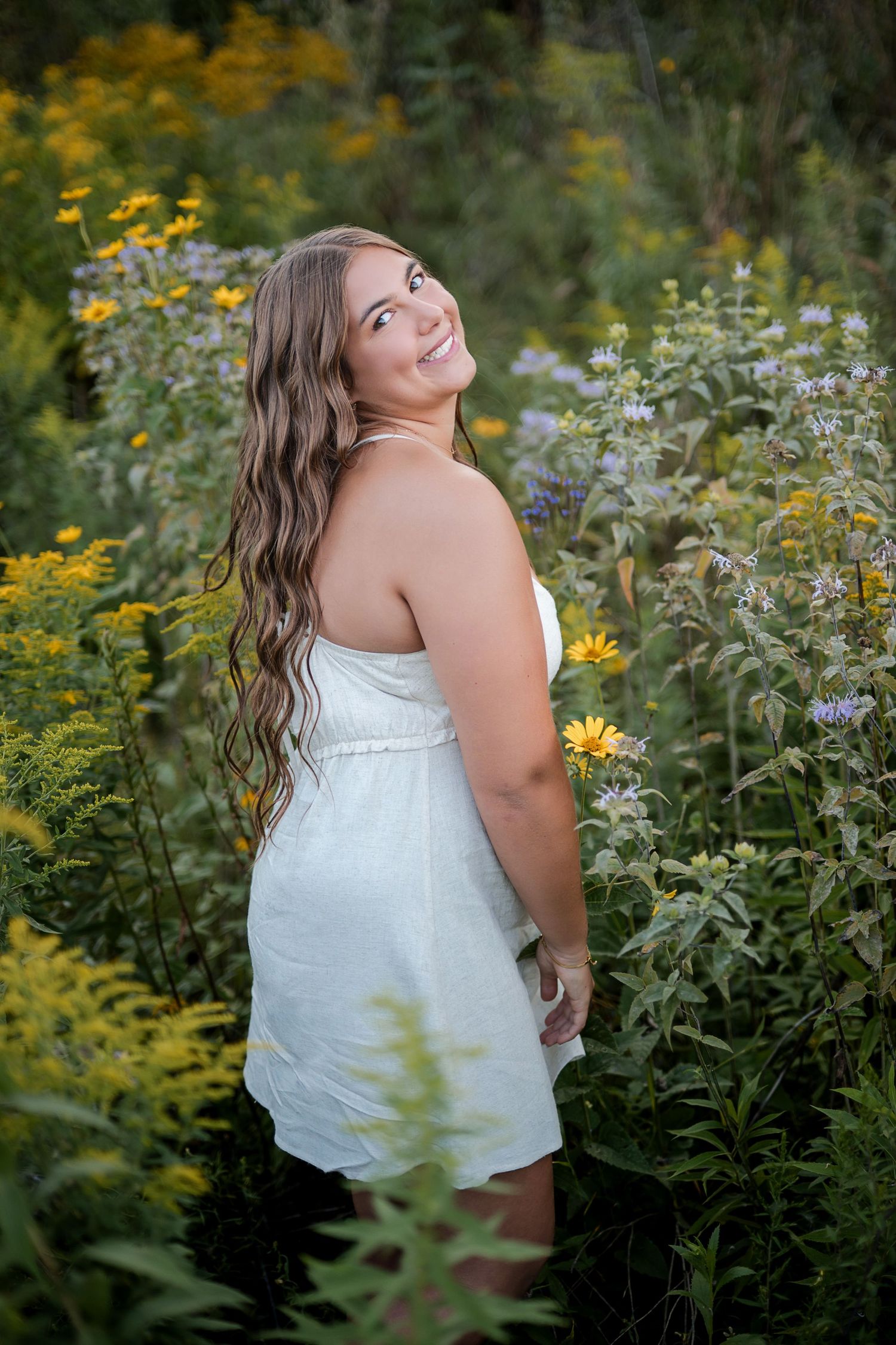 Young woman in white dress smiling among wildflowers and yellow blooms in a lush garden setting.