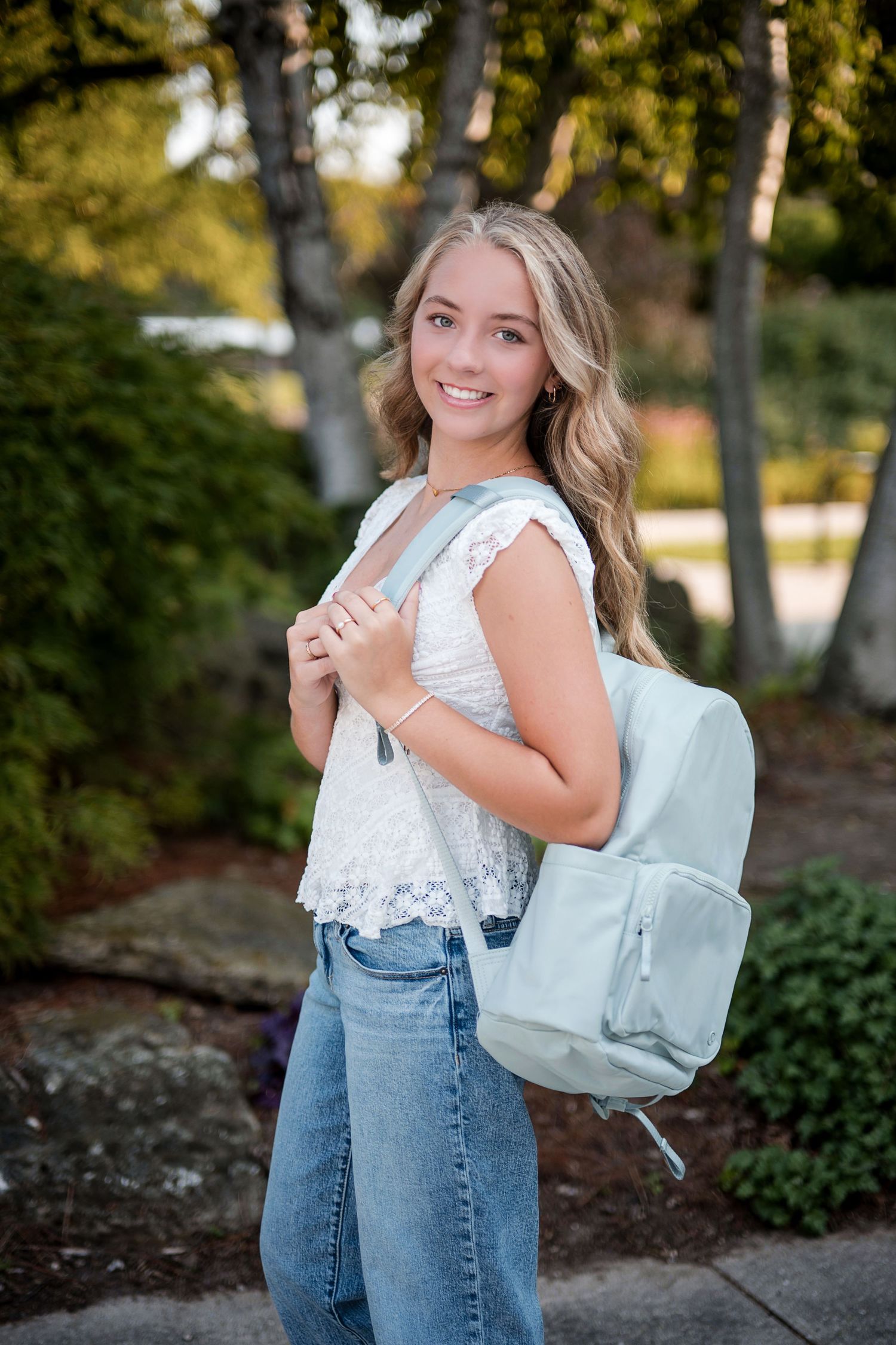 Smiling teenage girl with long blonde hair wearing a white lace top and a mint green backpack in a leafy park.