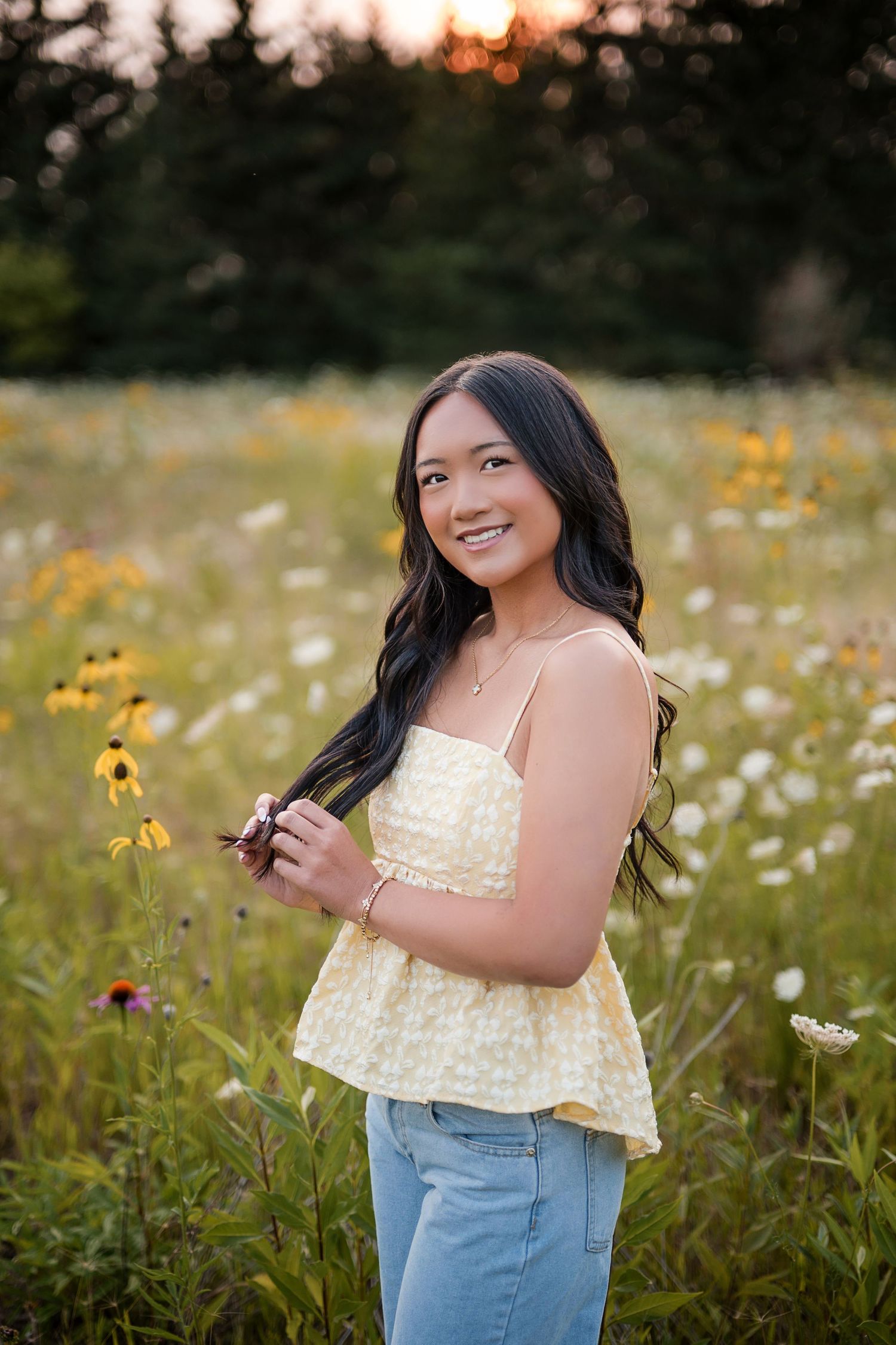 Young woman smiling in a wildflower field at sunset, wearing a yellow top and jeans, holding flowers.
