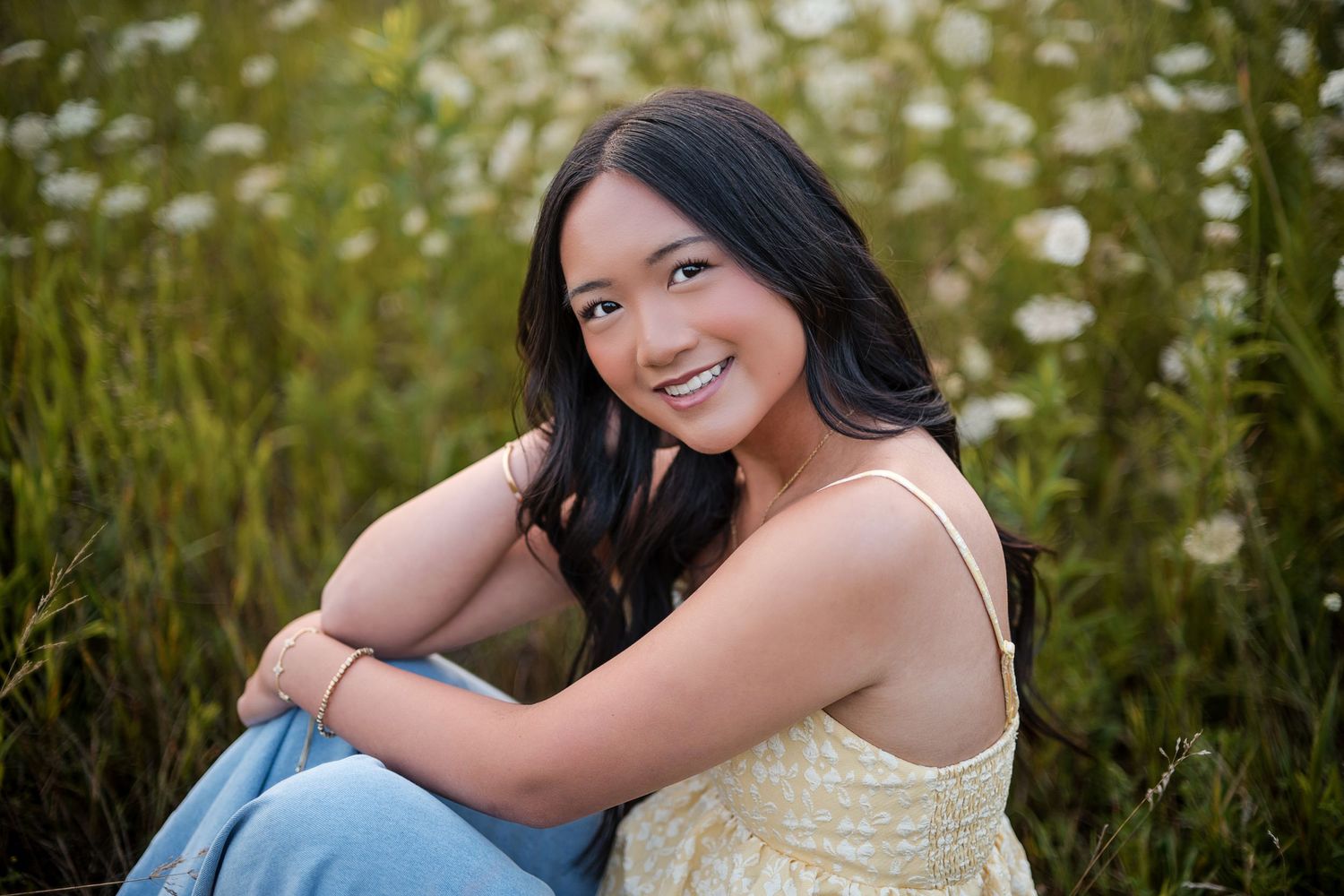 Young woman smiling in a wildflower field, wearing a soft yellow floral top and jeans, seated outdoors in natural light.