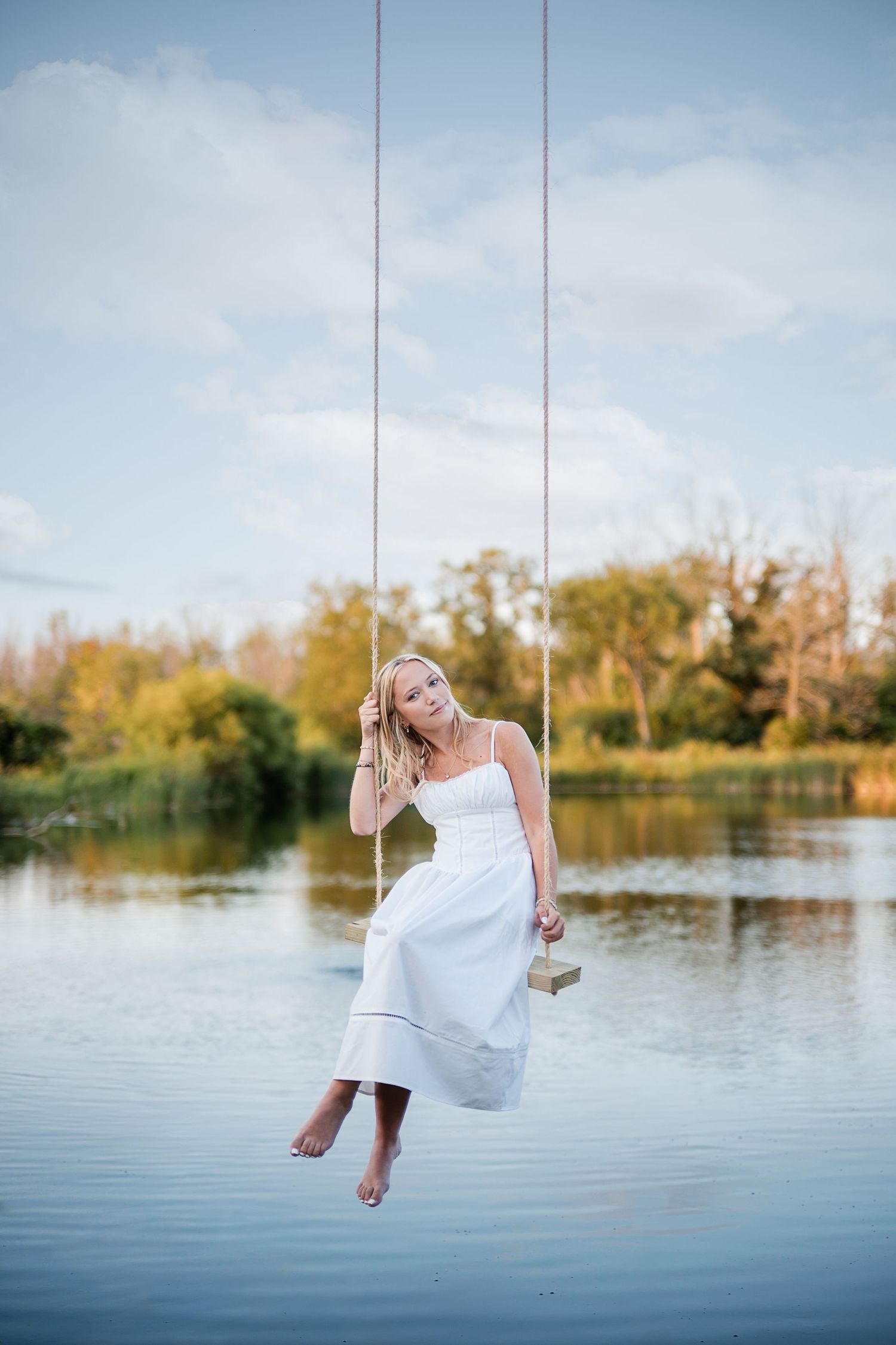 Barefoot woman in a white dress sits on a rope swing over a calm lake with autumn trees in the background.