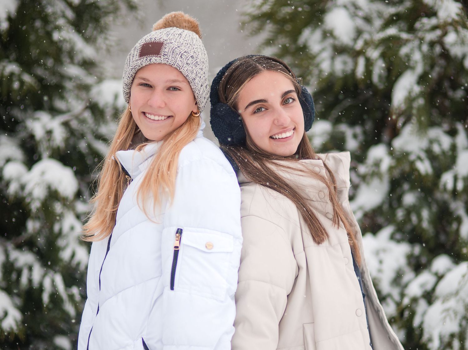 Two smiling young women in winter coats, a knit beanie and earmuffs, posing together in front of snow-covered evergreen trees.