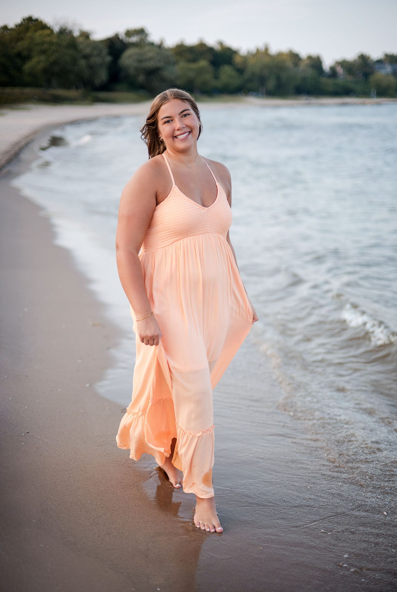 Young woman in a peach maxi dress smiling while walking barefoot along a sandy beach at golden hour.