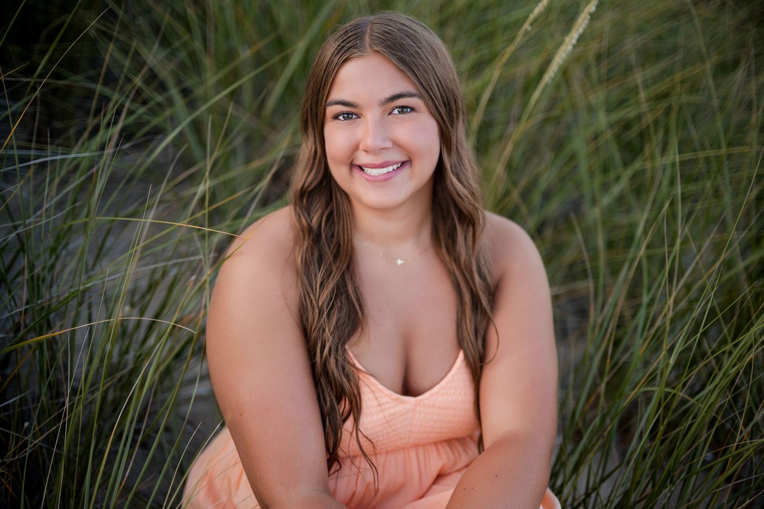 Smiling young woman with long brown hair wearing a peach dress, sitting outdoors among tall green beach grasses.