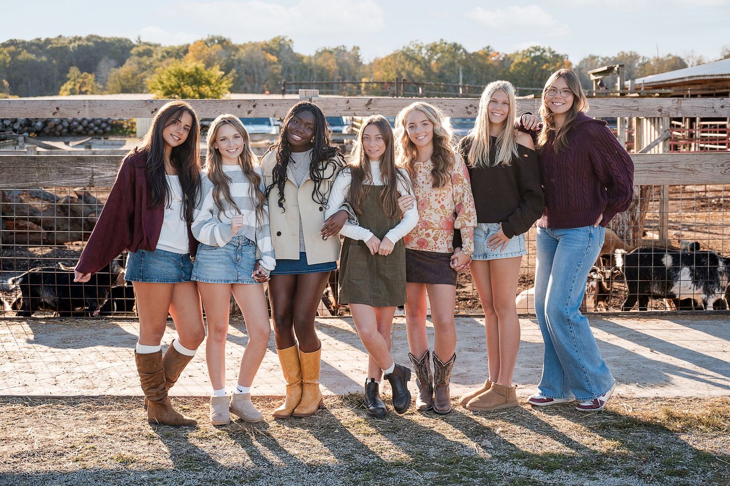Group of seven smiling girls in casual fall outfits and boots pose together by a wooden fence at golden hour.