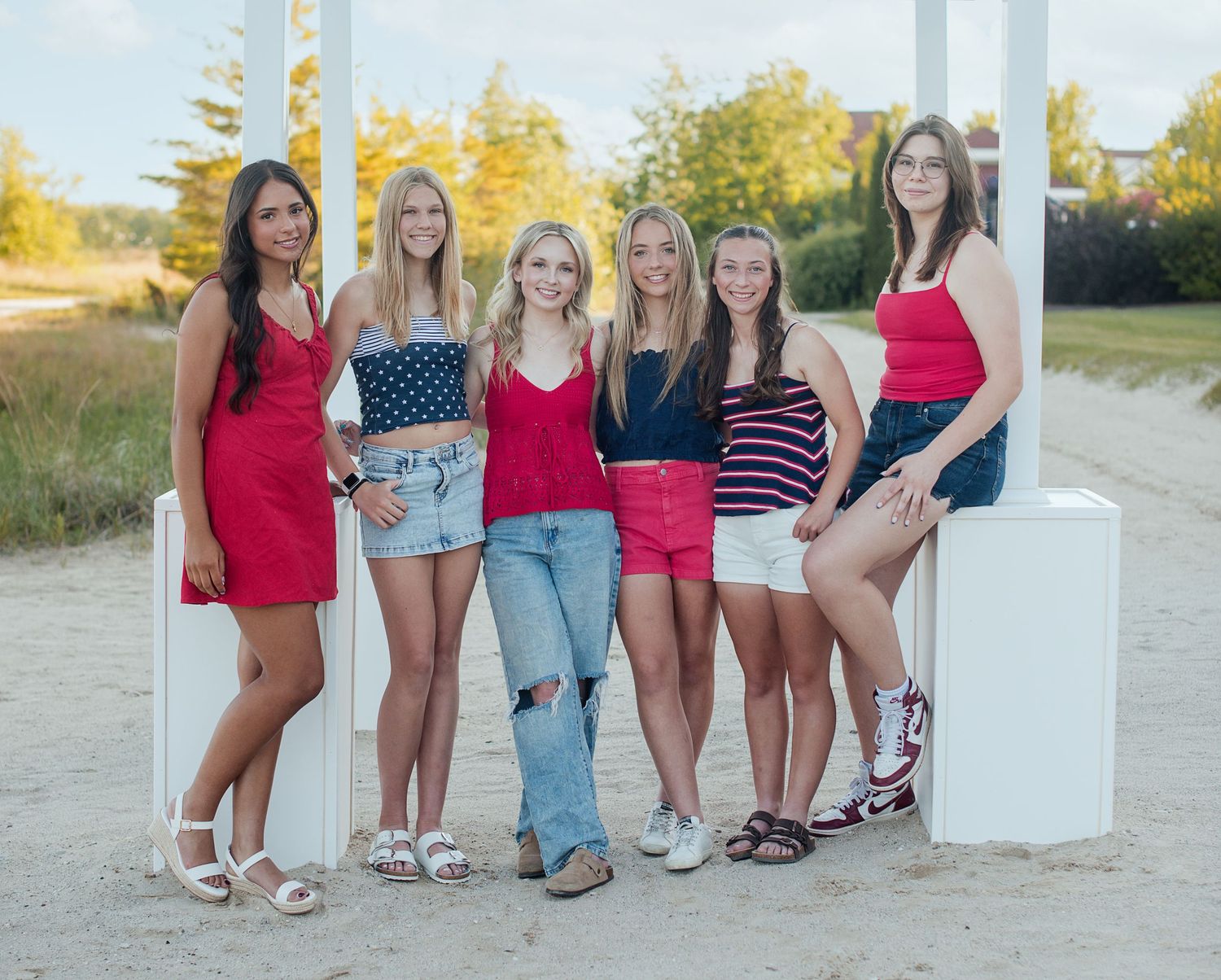 Six smiling teen girls in red, white and blue summer outfits pose together on white pedestals on a sandy beach path.