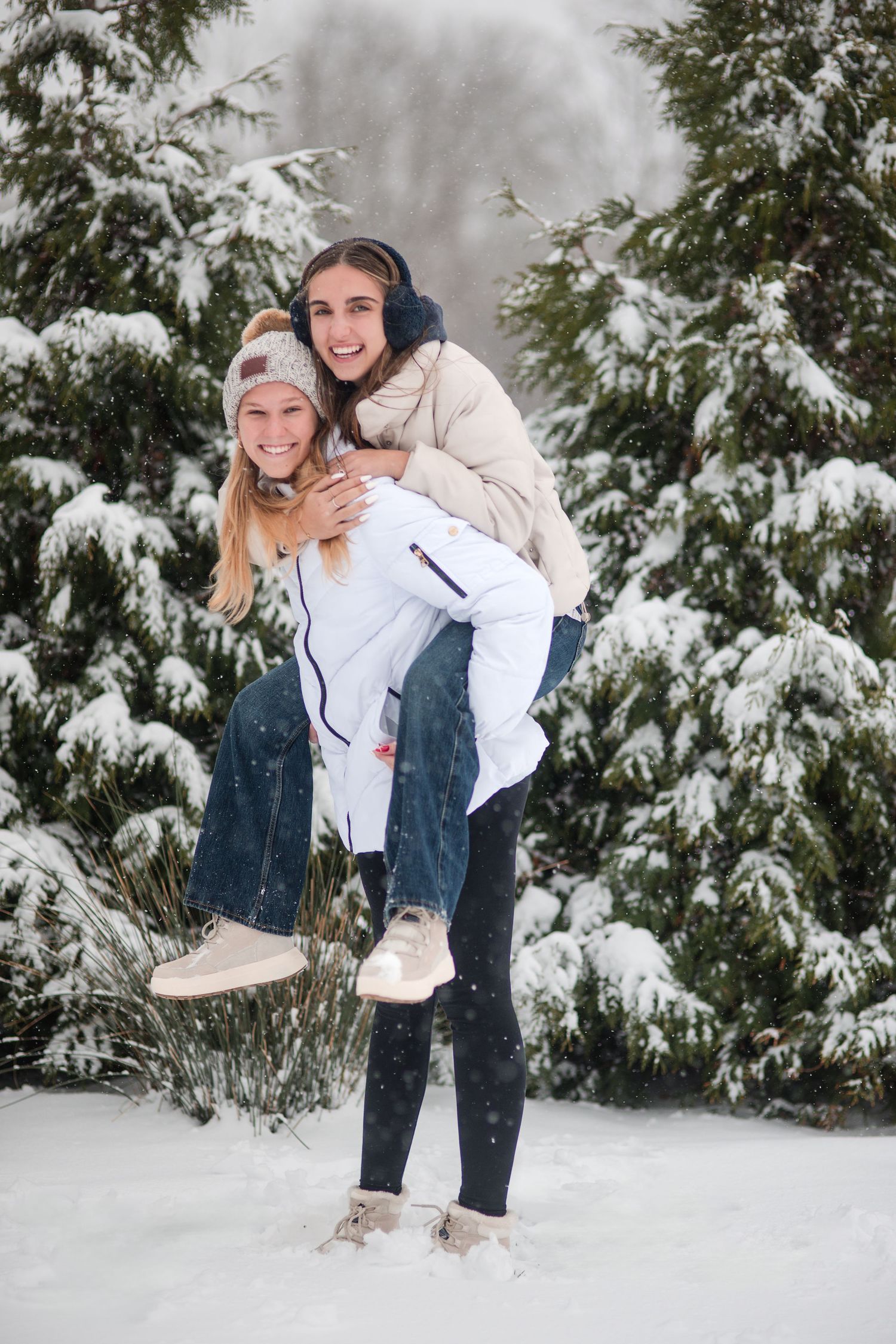 Smiling girl giving her friend a piggyback ride in winter gear in front of snow-covered evergreen trees.