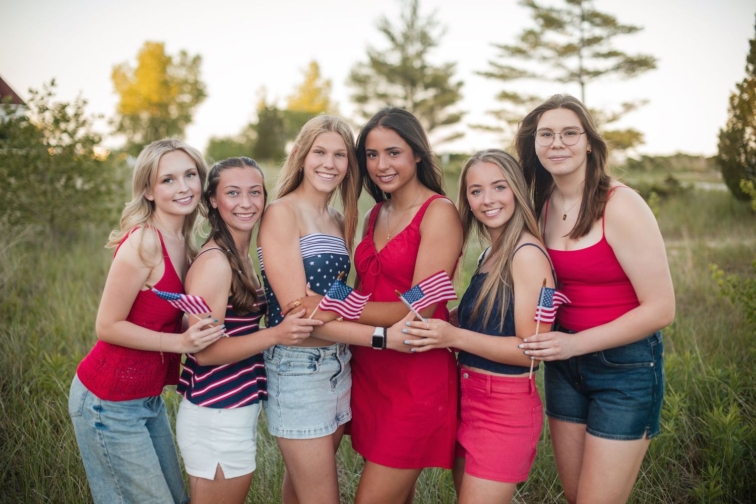 Six smiling friends in patriotic red, white, and blue outfits posing with small American flags in a grassy field at sunset.