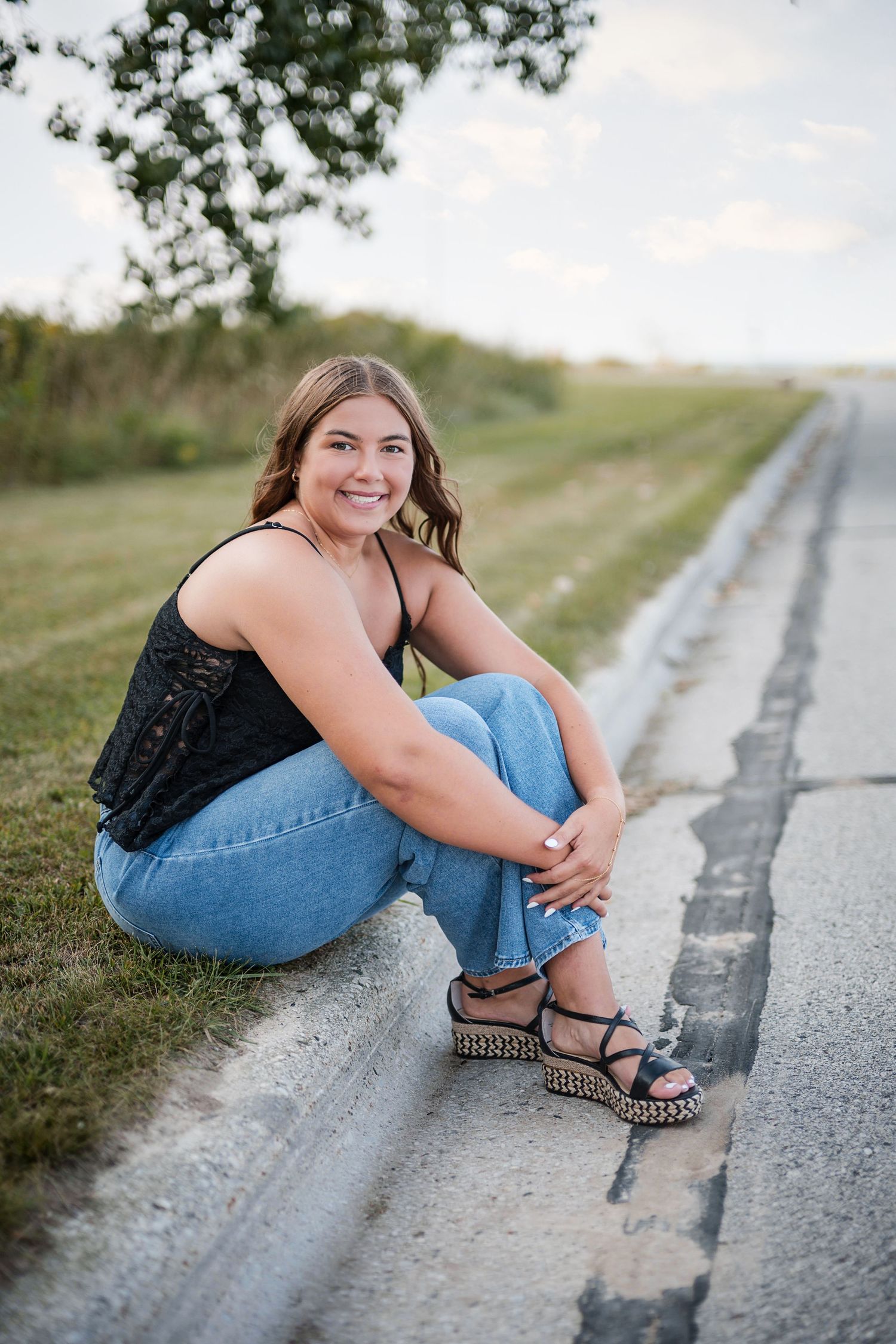Young woman in black top and jeans sits smiling on a roadside curb wearing wedge sandals on a sunny day.