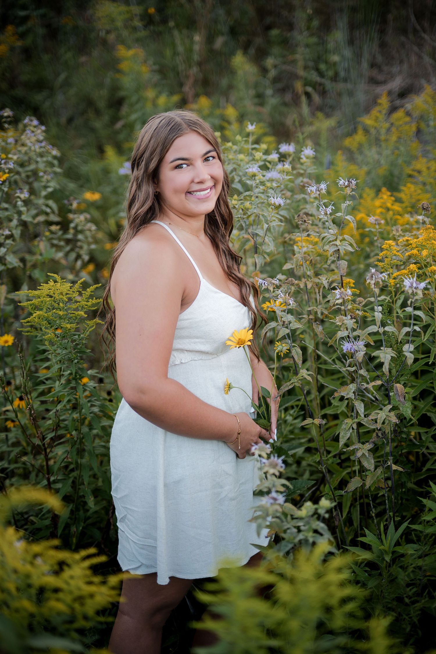 Young woman in white sundress smiling among wildflowers, holding a yellow bloom in a lush garden setting.