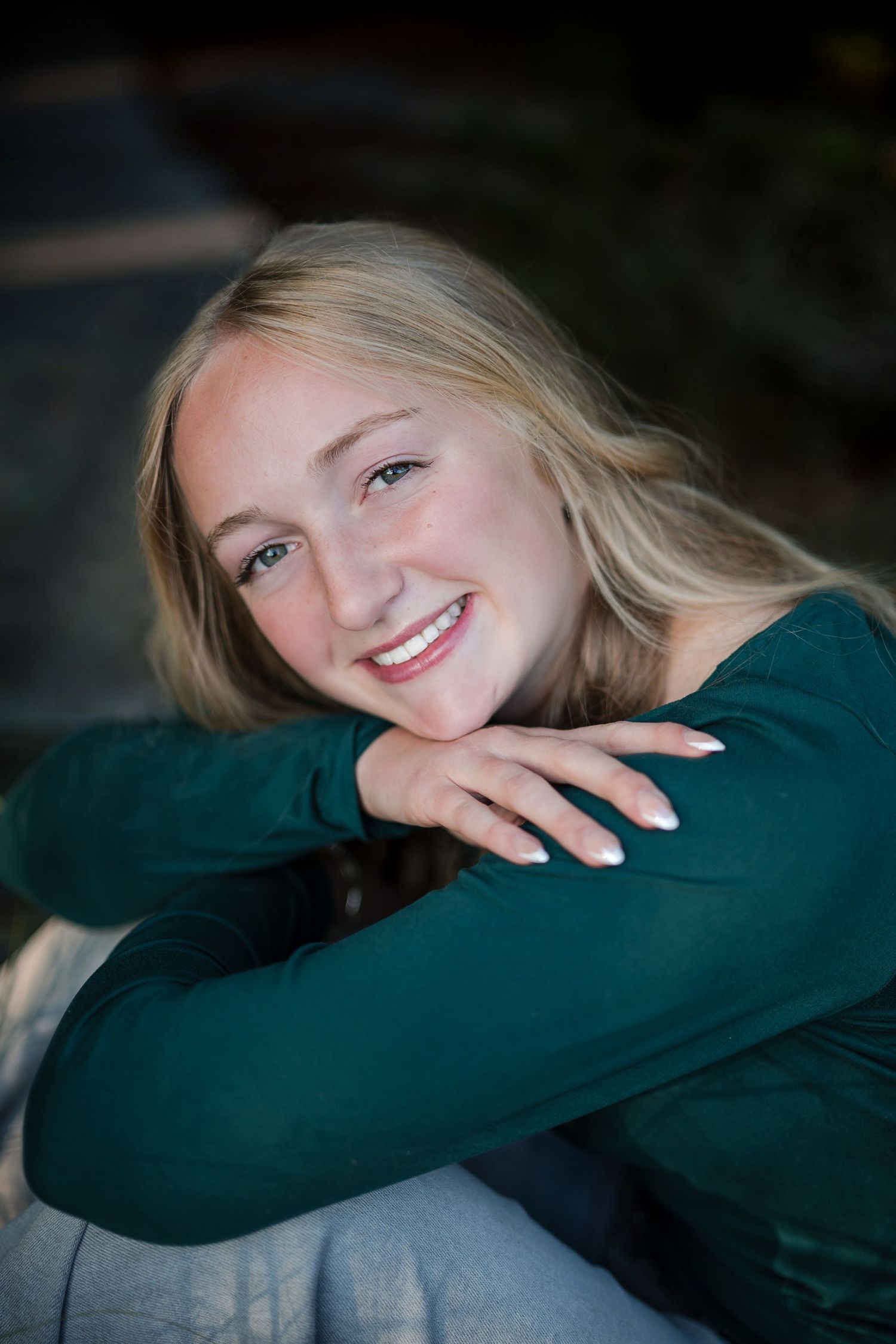 Smiling teenage girl with long blonde hair in a green top rests her chin on her folded arms in a senior portrait.