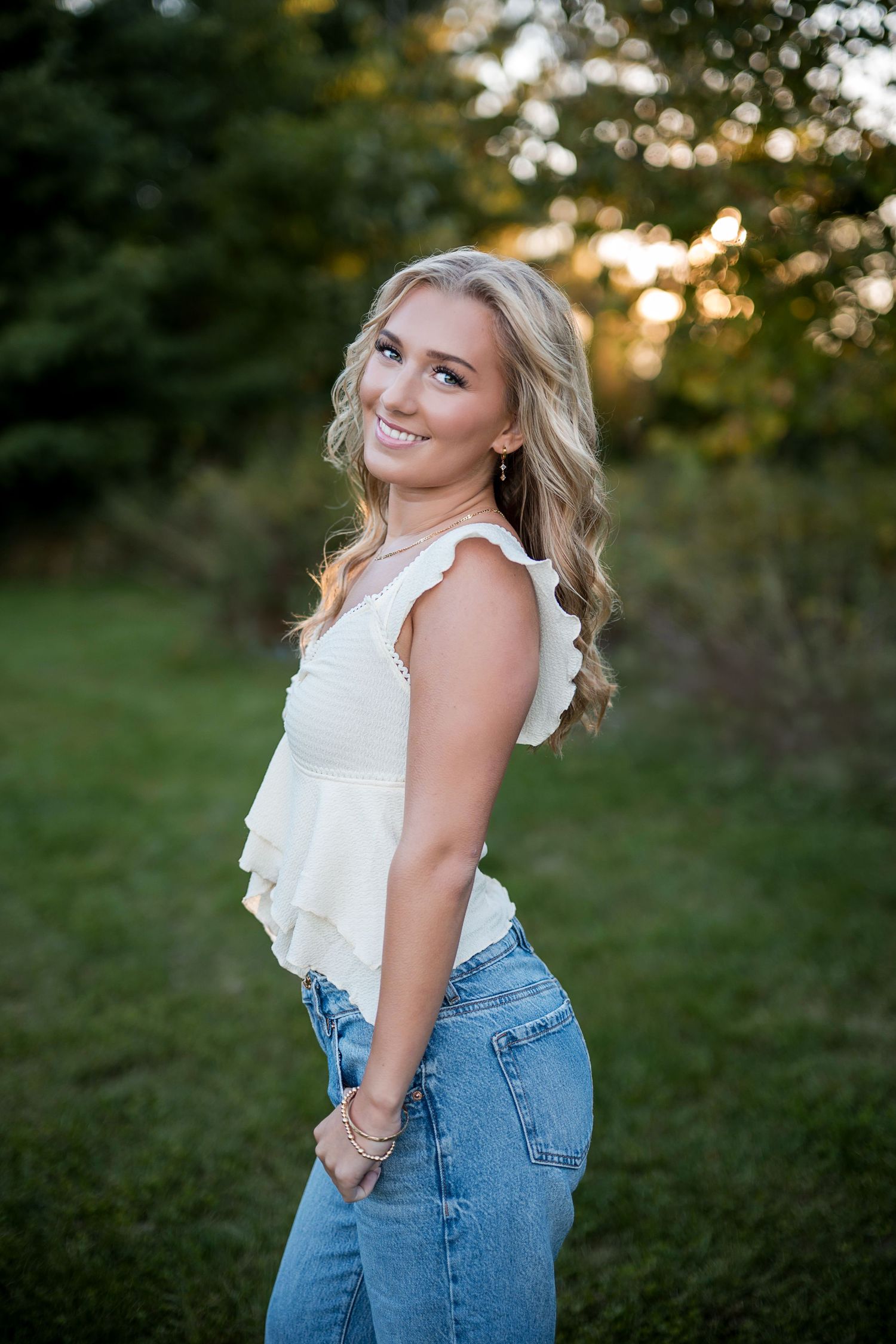 Blonde young woman in a cream ruffled top and blue jeans smiles over her shoulder in a green park during golden hour.