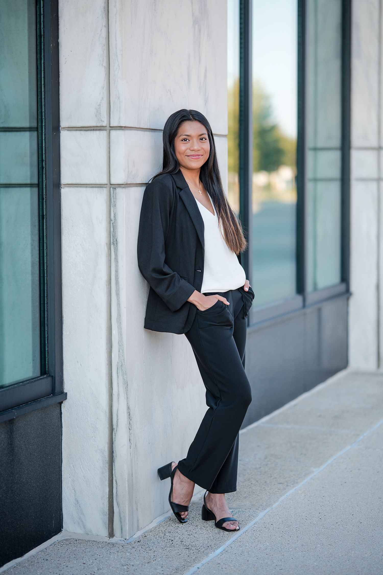 Young woman in black blazer and pants leans against marble wall outside modern building.
