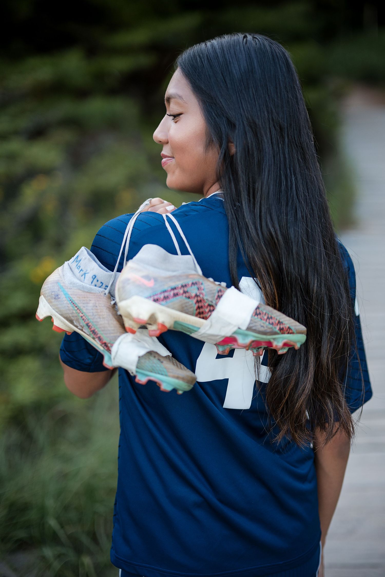 Young girl in blue jersey holds colorful Nike cleats over her shoulder, smiling outdoors near green trees.