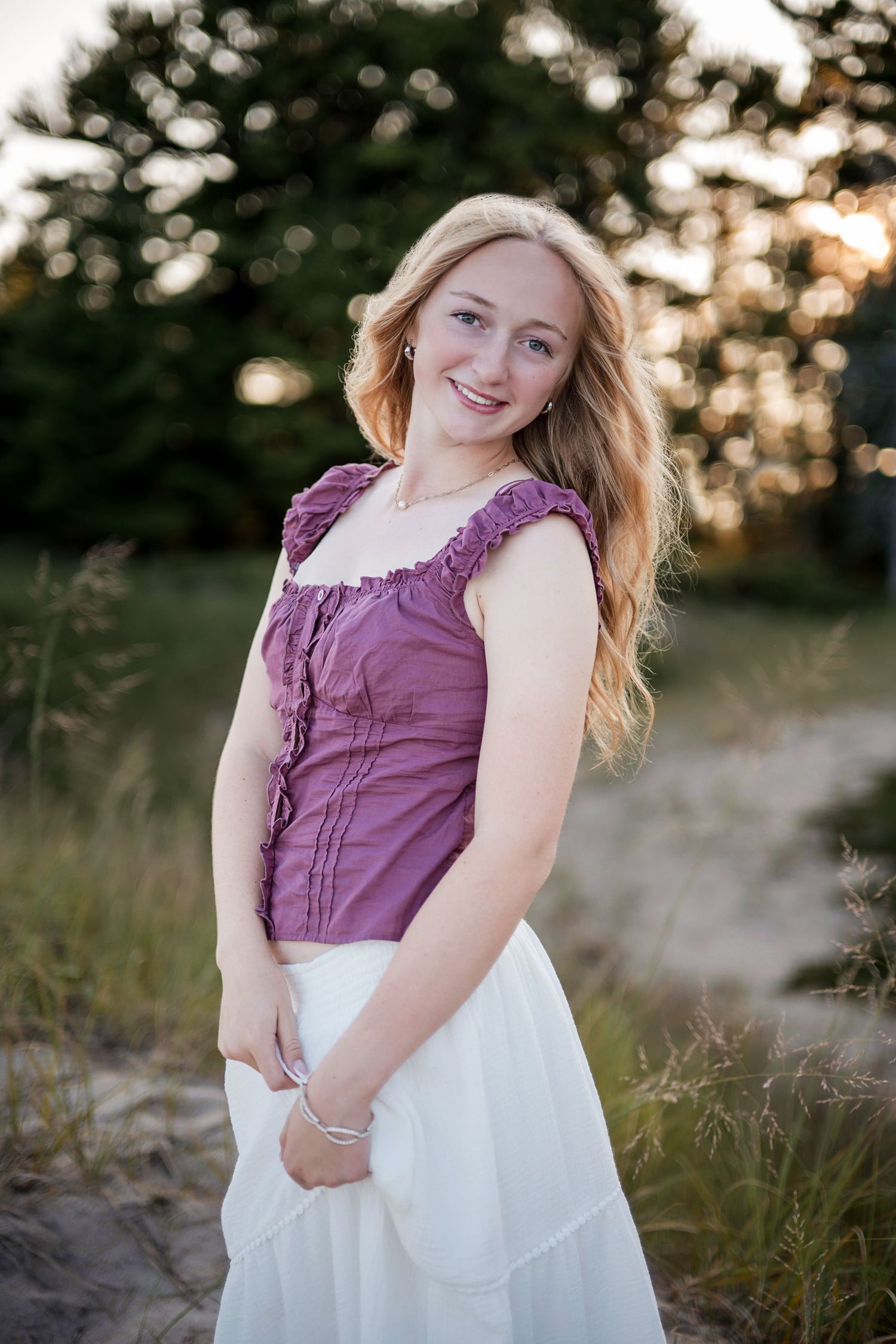 Smiling young blonde woman in purple top and white skirt posing outdoors at golden hour.