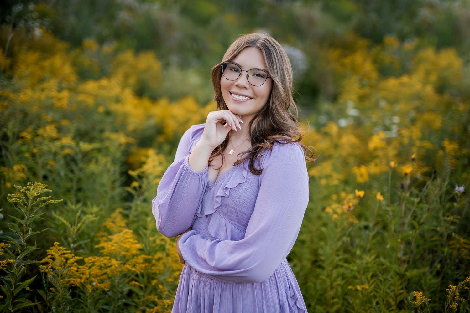 Smiling young woman in glasses and a lavender dress poses in a field of yellow wildflowers during an outdoor portrait session.