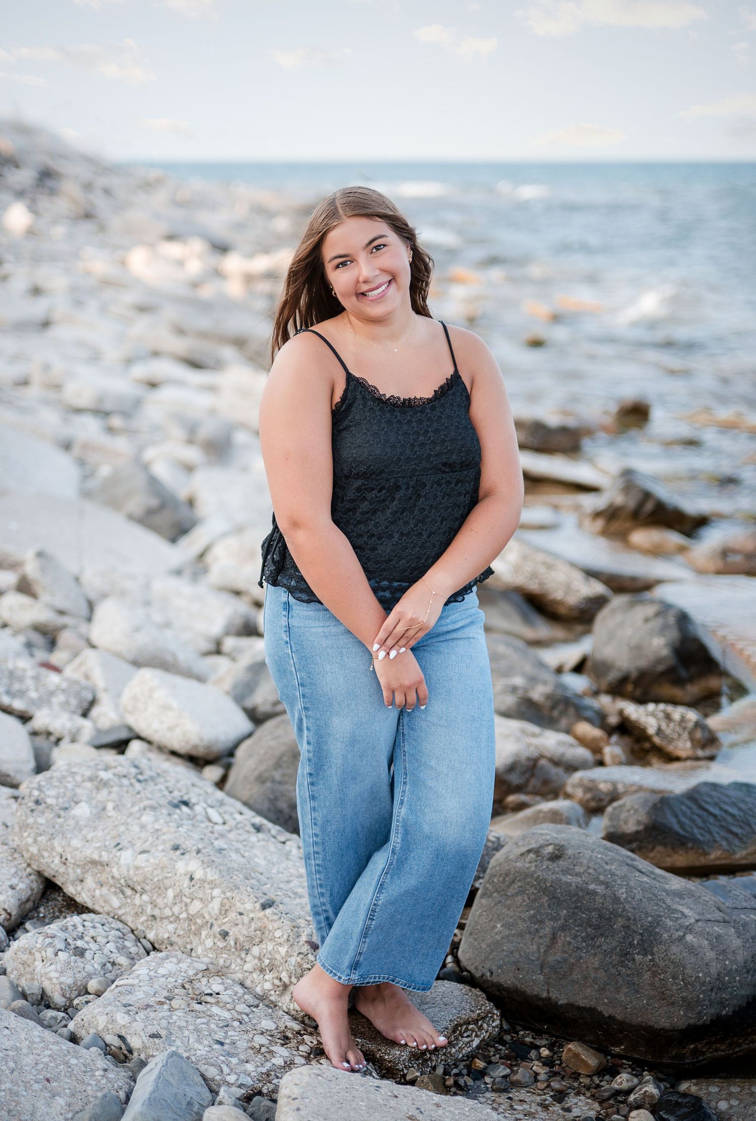 Young woman in black tank top and jeans smiling on rocky Lake Michigan shoreline during golden hour.