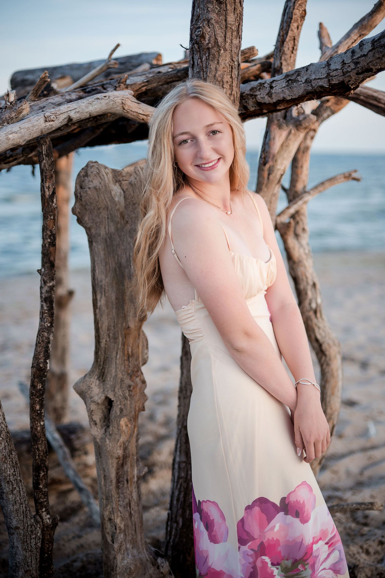 Smiling blonde woman in floral maxi dress posing by driftwood structure on a sandy beach at dusk.