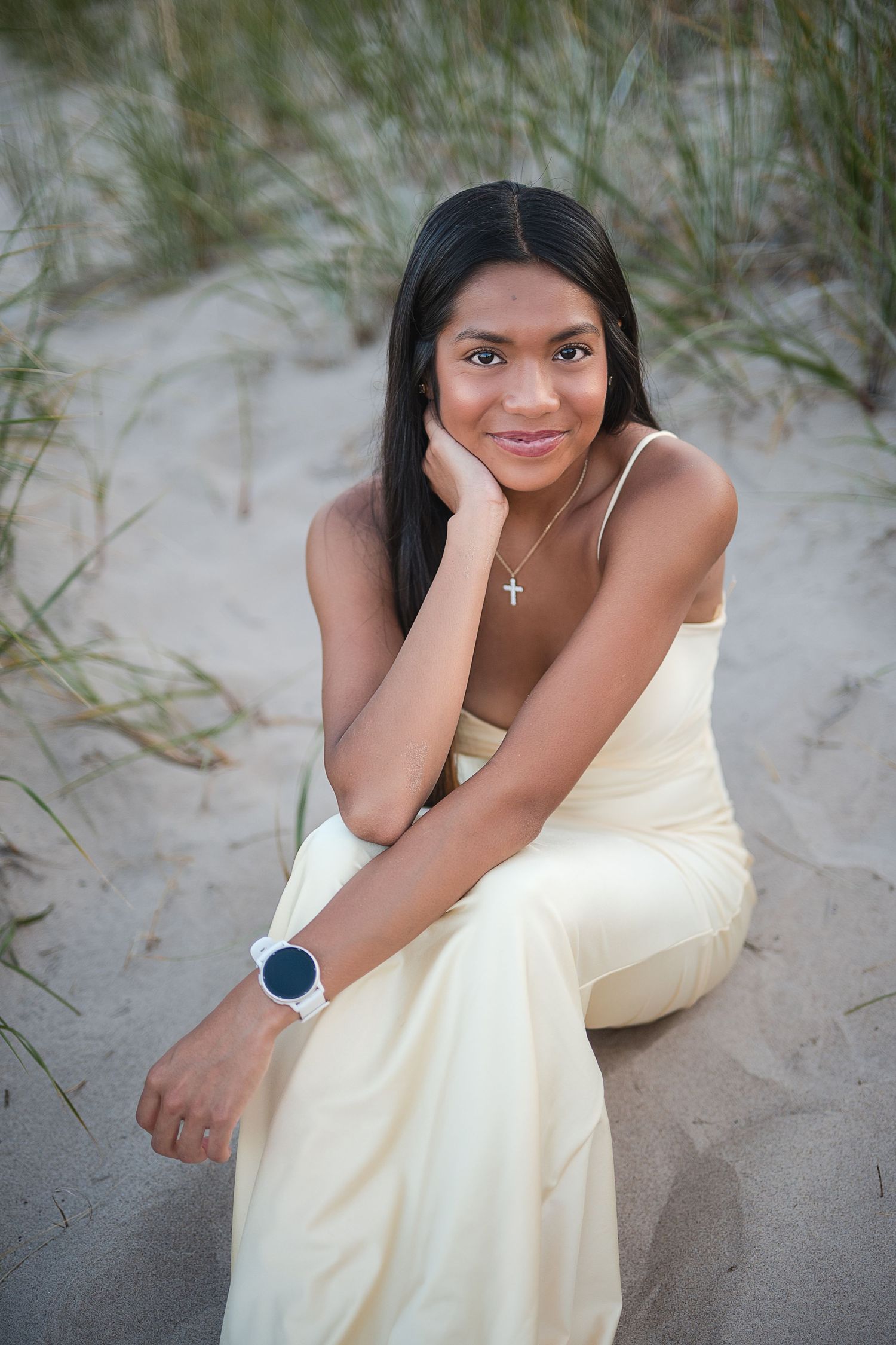 Young woman in cream dress sitting on sandy beach dunes, wearing a cross necklace and white watch.