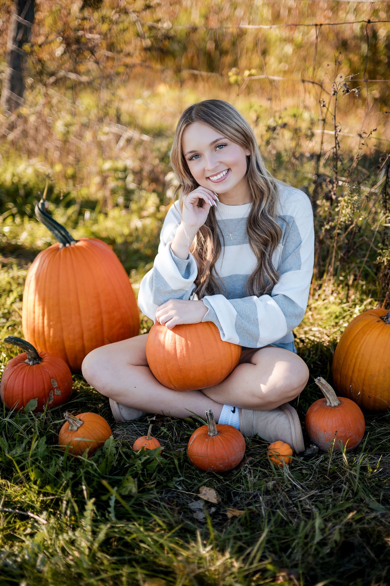 Smiling teen girl in a striped sweater sits on grass surrounded by orange pumpkins in a sunny autumn field.