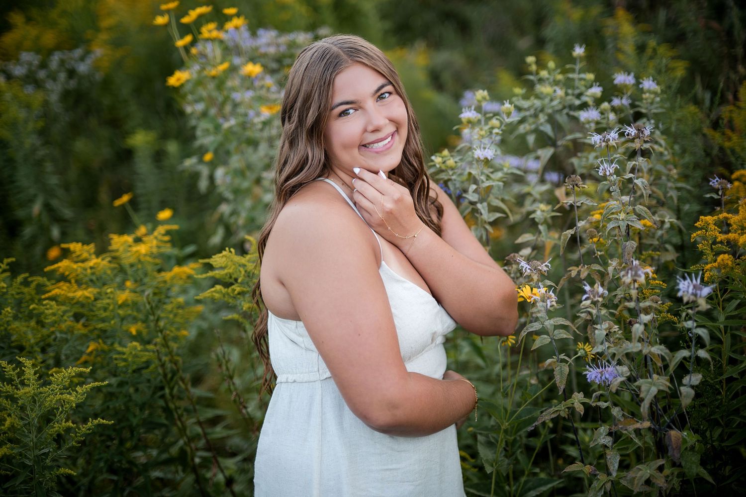 Young woman in white dress smiling among wildflowers, golden hour senior portrait photography outdoors.