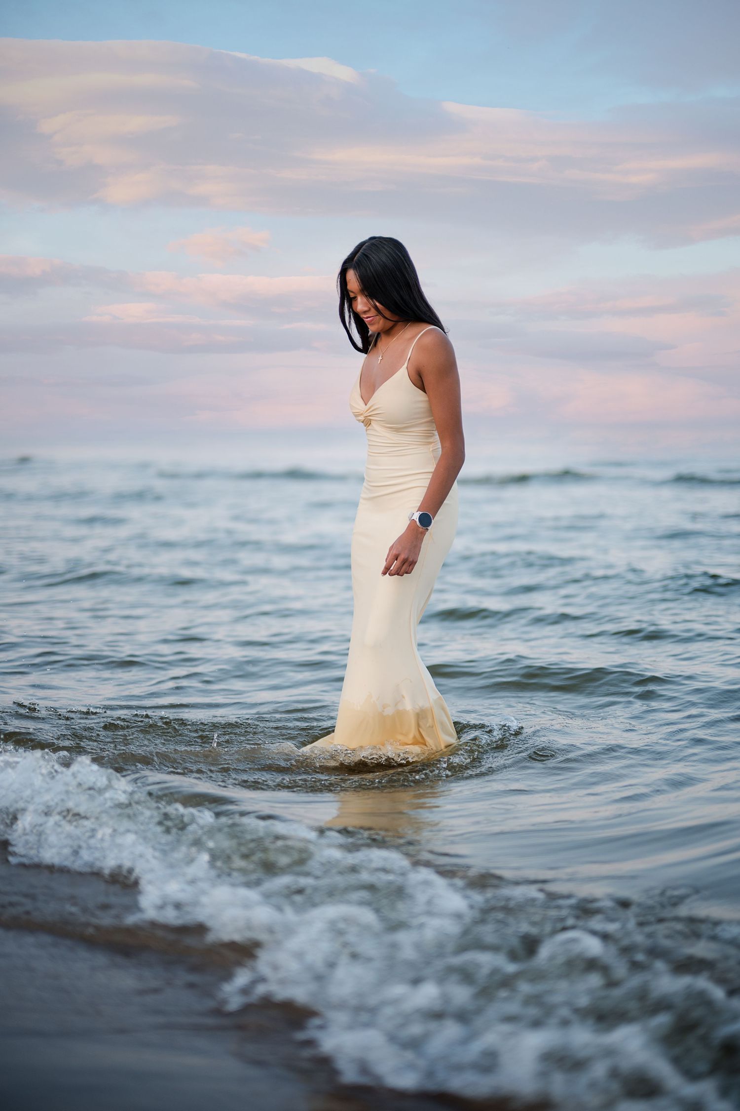 Woman in elegant white gown standing in ocean waves at sunset, looking down at the water.