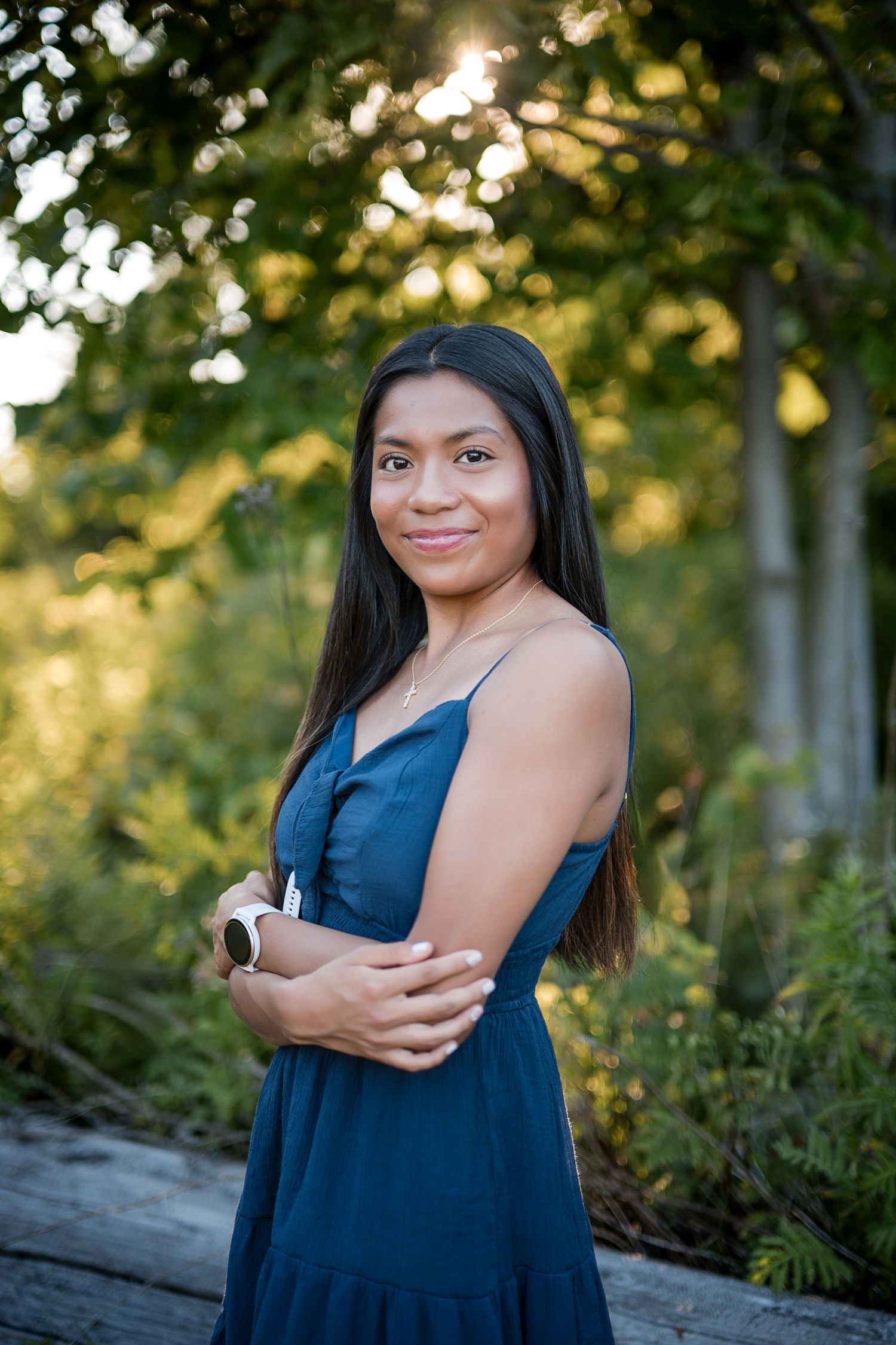 Young woman in teal dress poses outdoors with golden sunlight filtering through green trees behind her.
