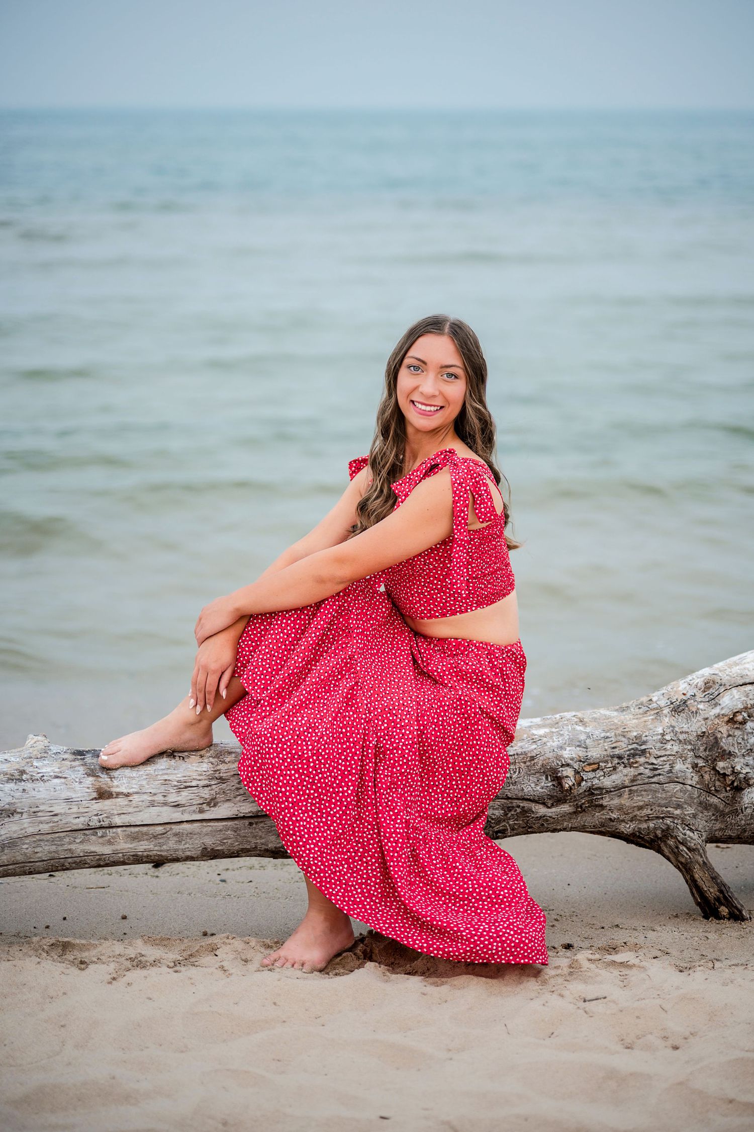 Smiling young woman in a red polka dot two-piece outfit sits barefoot on a driftwood log on a sandy beach beside calm lake water.
