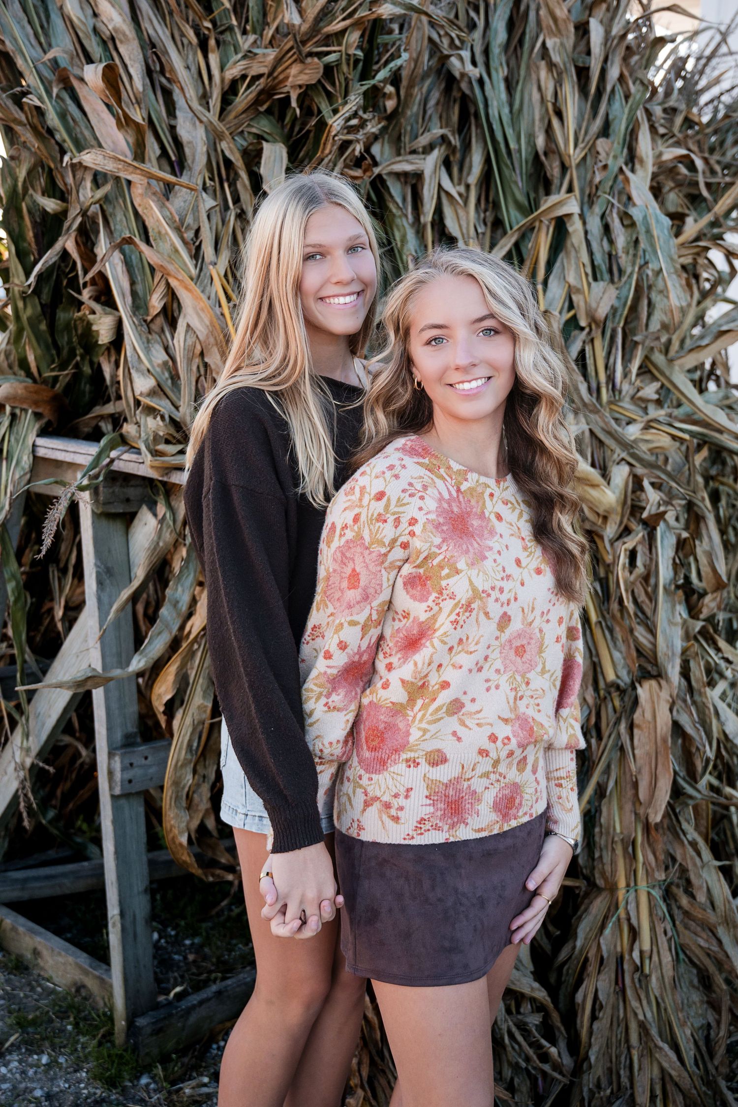 Two smiling girls stand arm in arm in front of a corn stalk display, one in a brown sweater and one in a floral top.