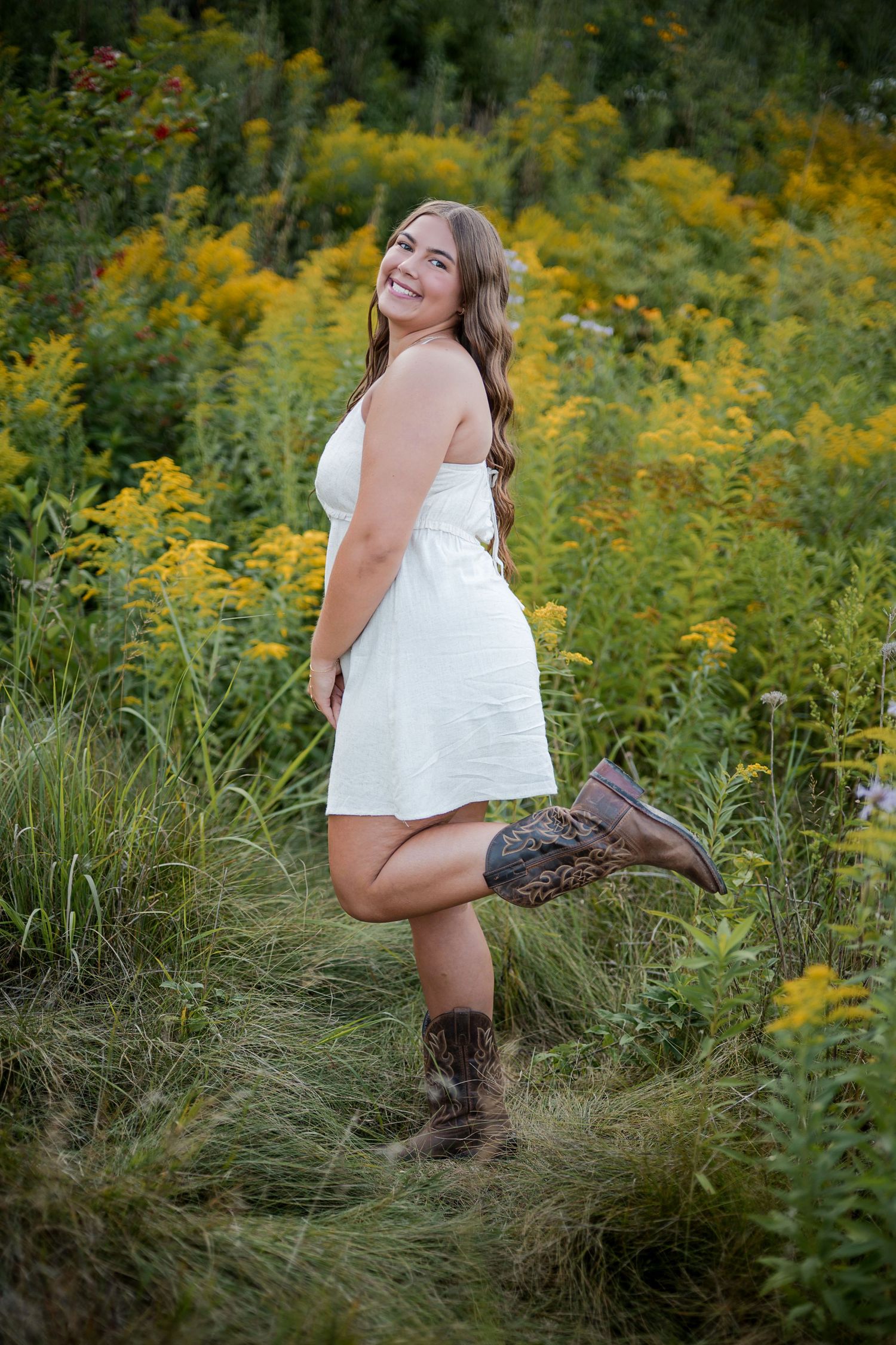 Young woman in white dress and cowboy boots smiling in a wildflower field.