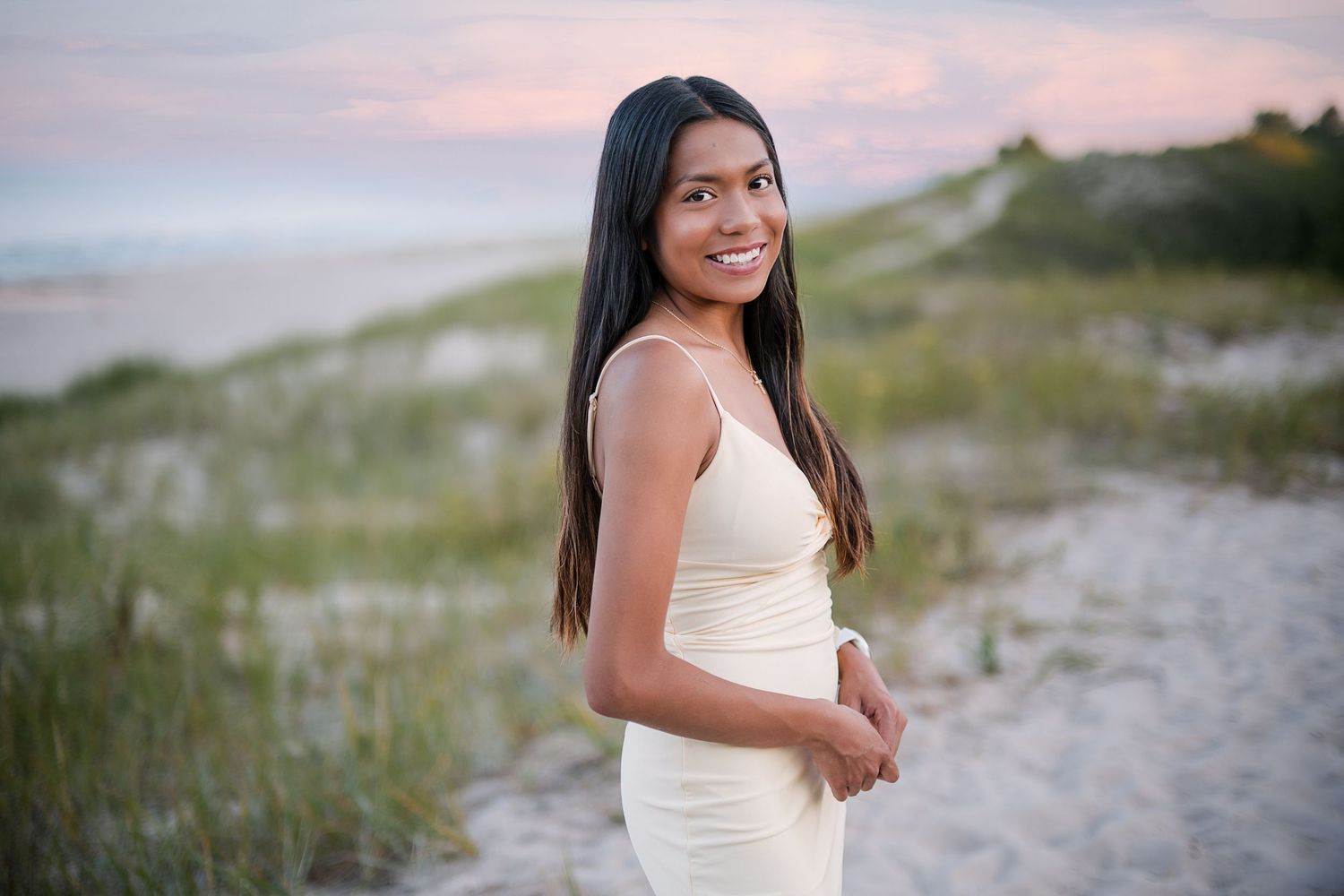 Young woman in cream dress smiling on sandy beach at sunset with dunes and pastel sky behind her.