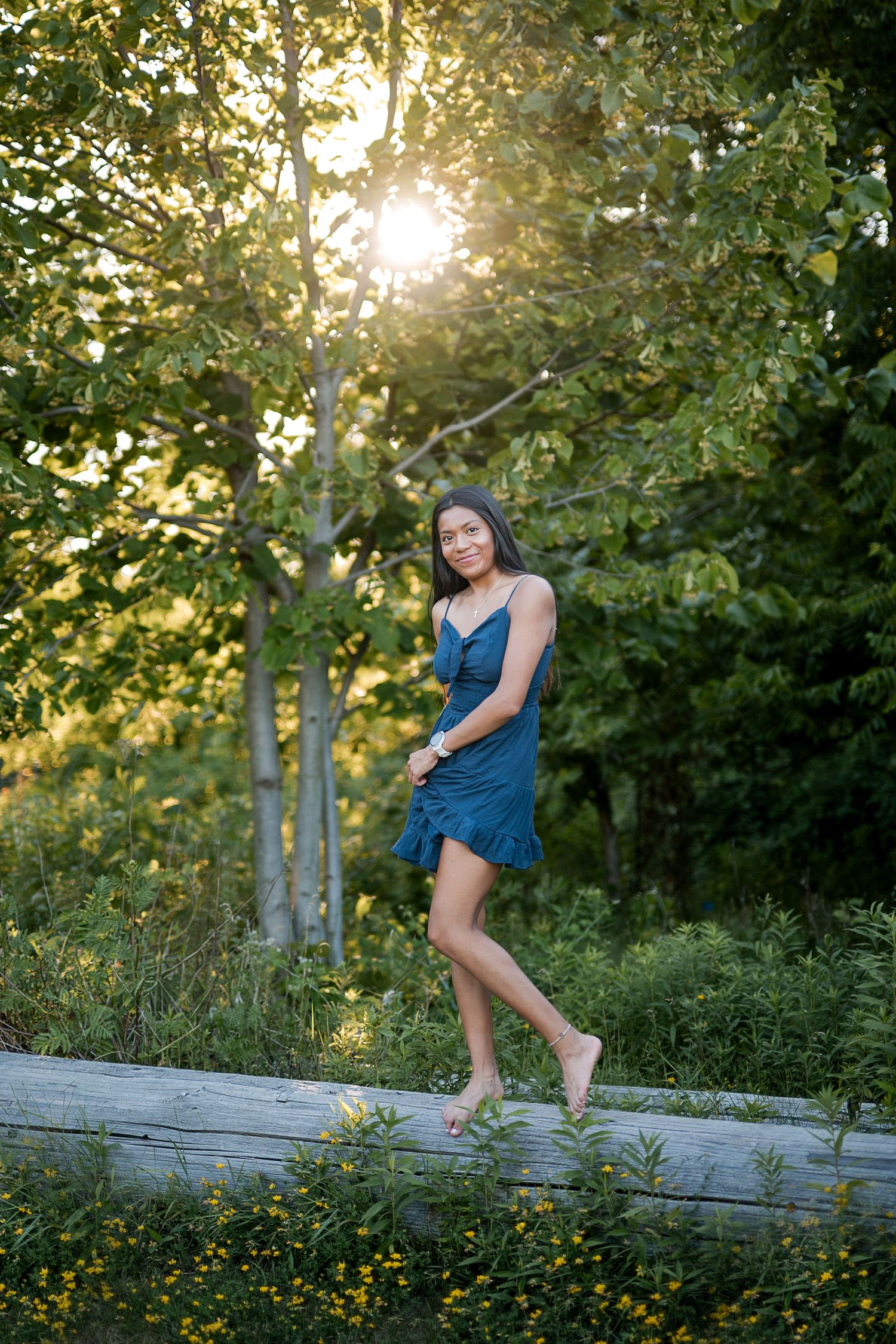 Young woman in teal dress standing barefoot on a log in a sunlit forest during golden hour.