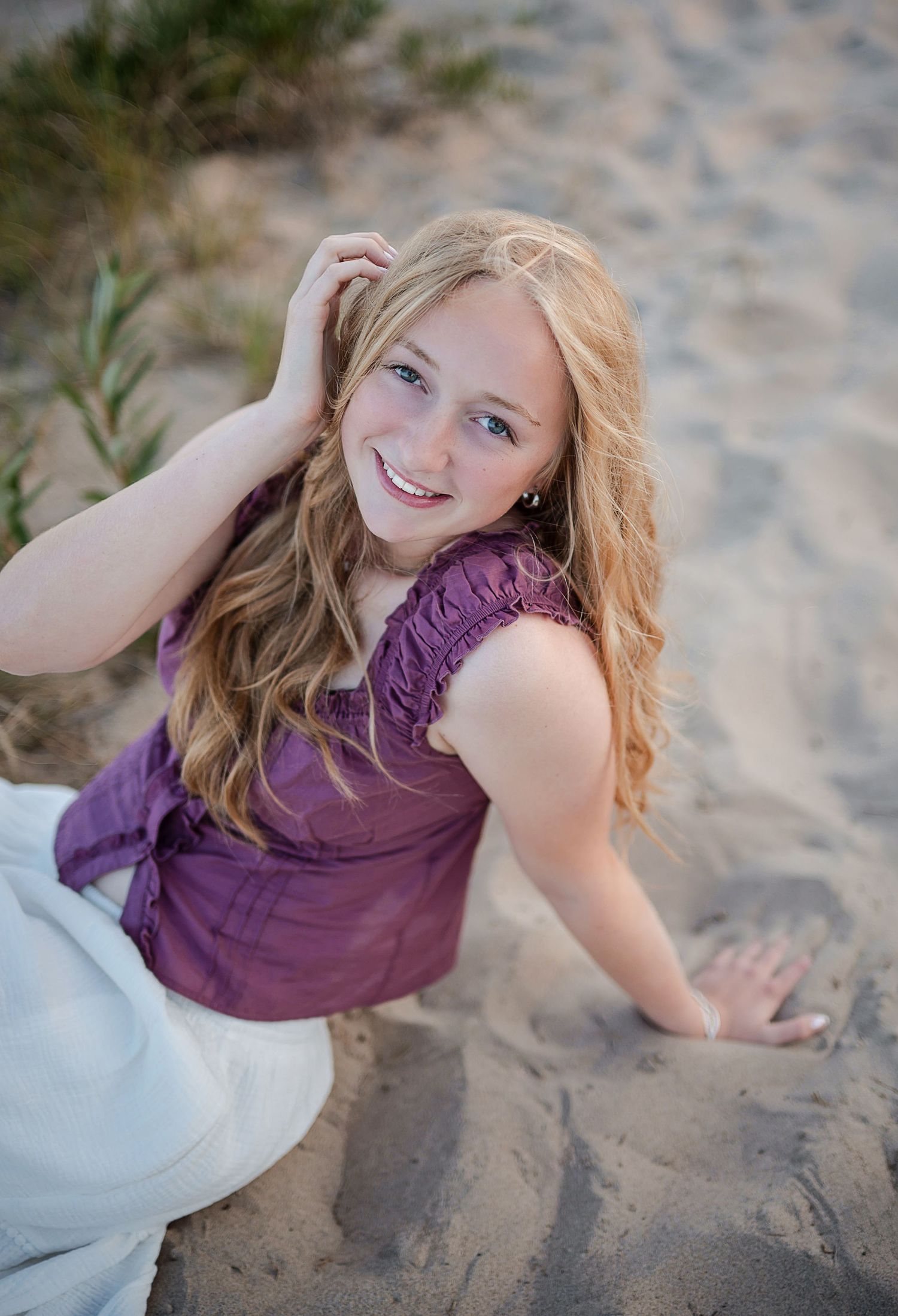 Young blonde woman in purple top sitting on sandy beach dunes, smiling and posing for senior portrait photo.