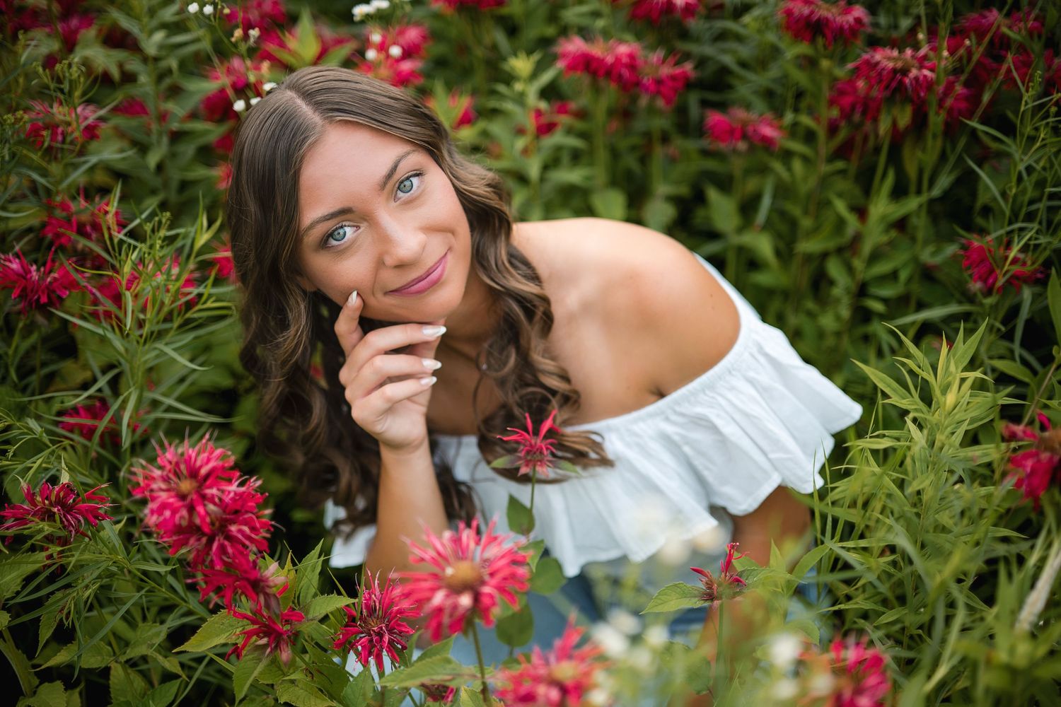 Smiling young woman with curly brown hair in white off-shoulder top surrounded by vibrant red flowers.