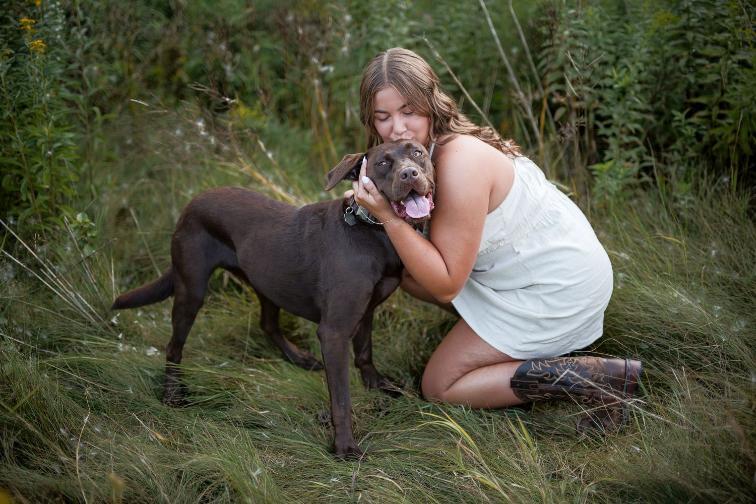 Young woman in white dress hugging a chocolate Labrador dog in a lush green field.