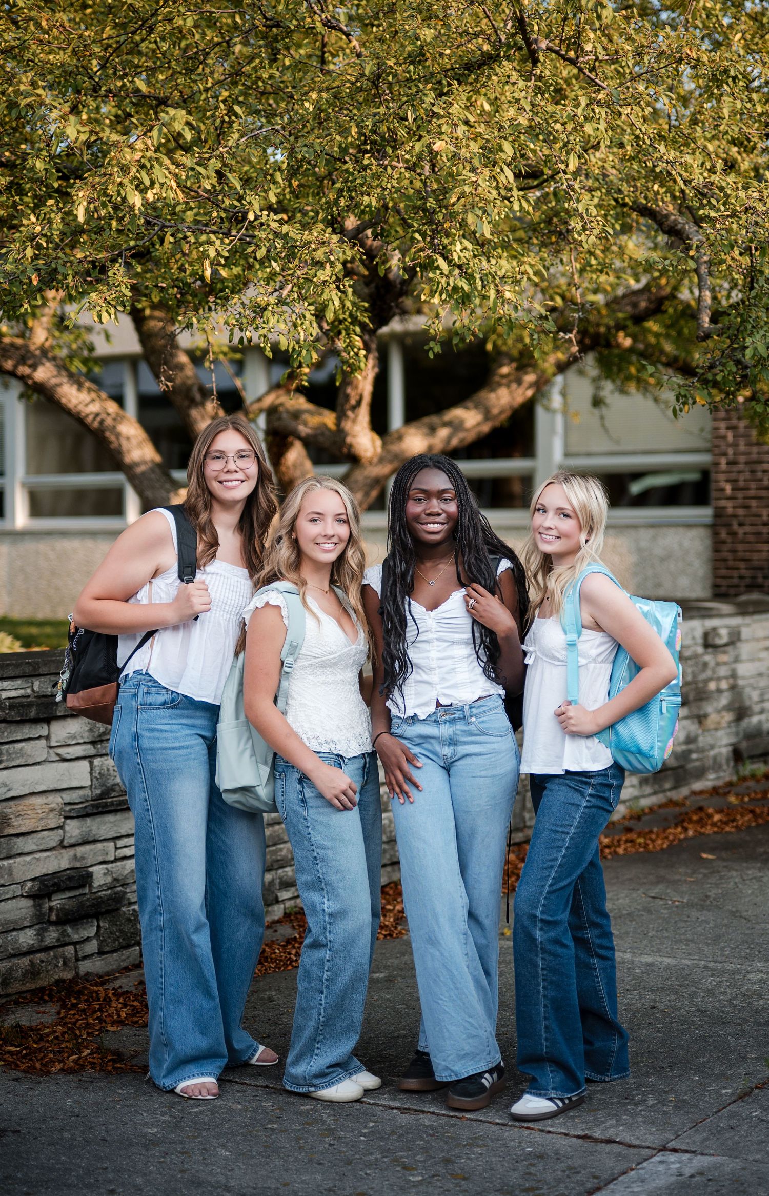 Four smiling girls in white tops and wide-leg jeans with backpacks pose under a leafy tree by a stone wall.