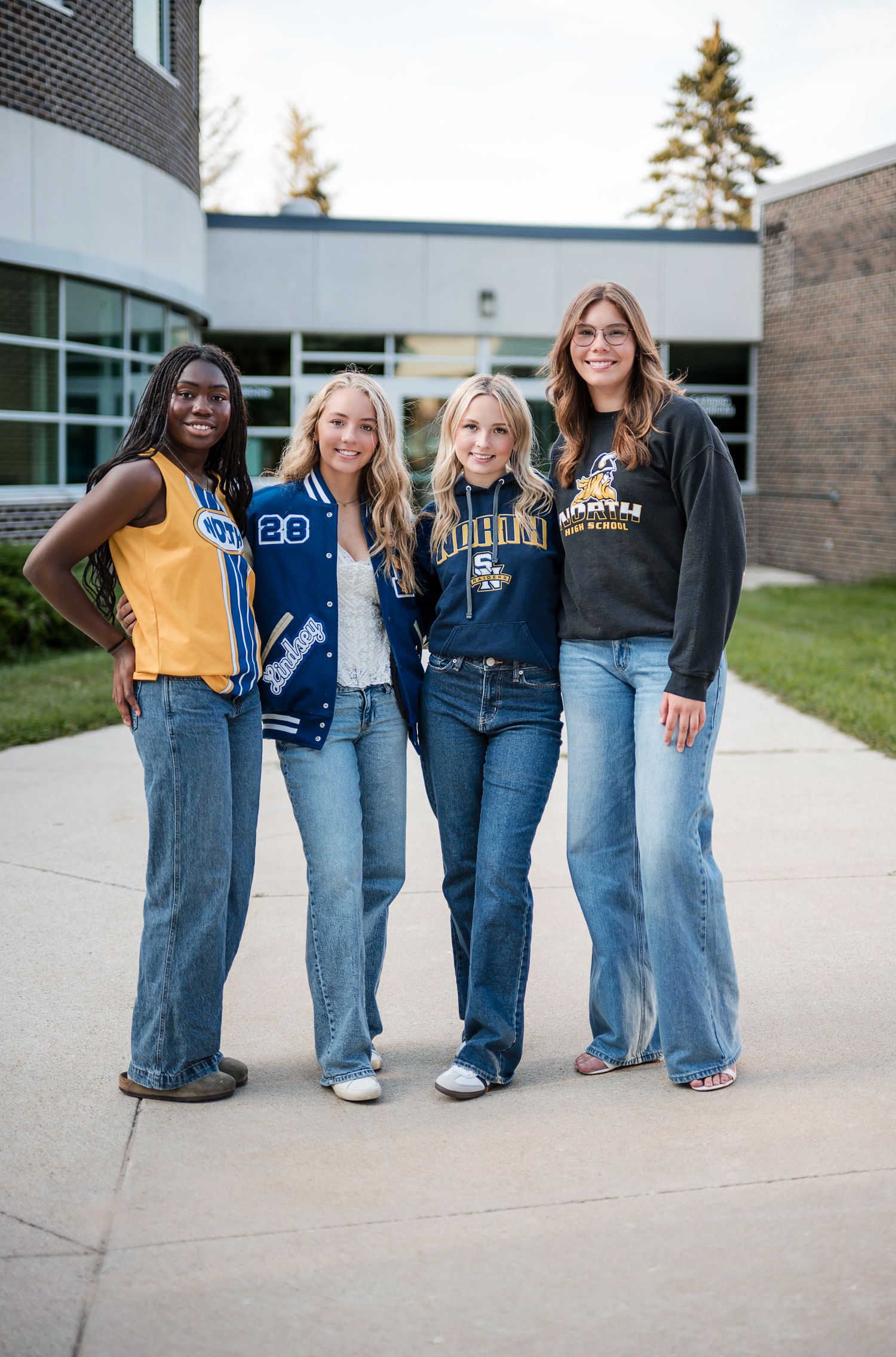 Four smiling friends in jeans and school spirit apparel posing together on a sidewalk outside a school building.