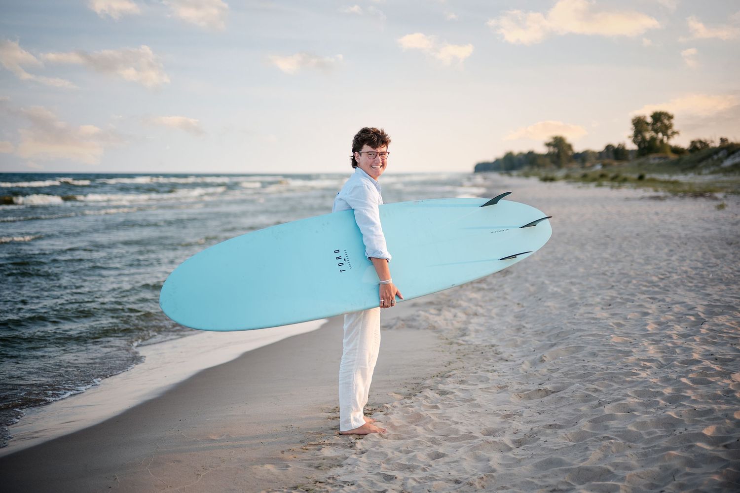 Young person in white clothing carries a light blue surfboard along a sandy beach beside gentle ocean waves at sunset.
