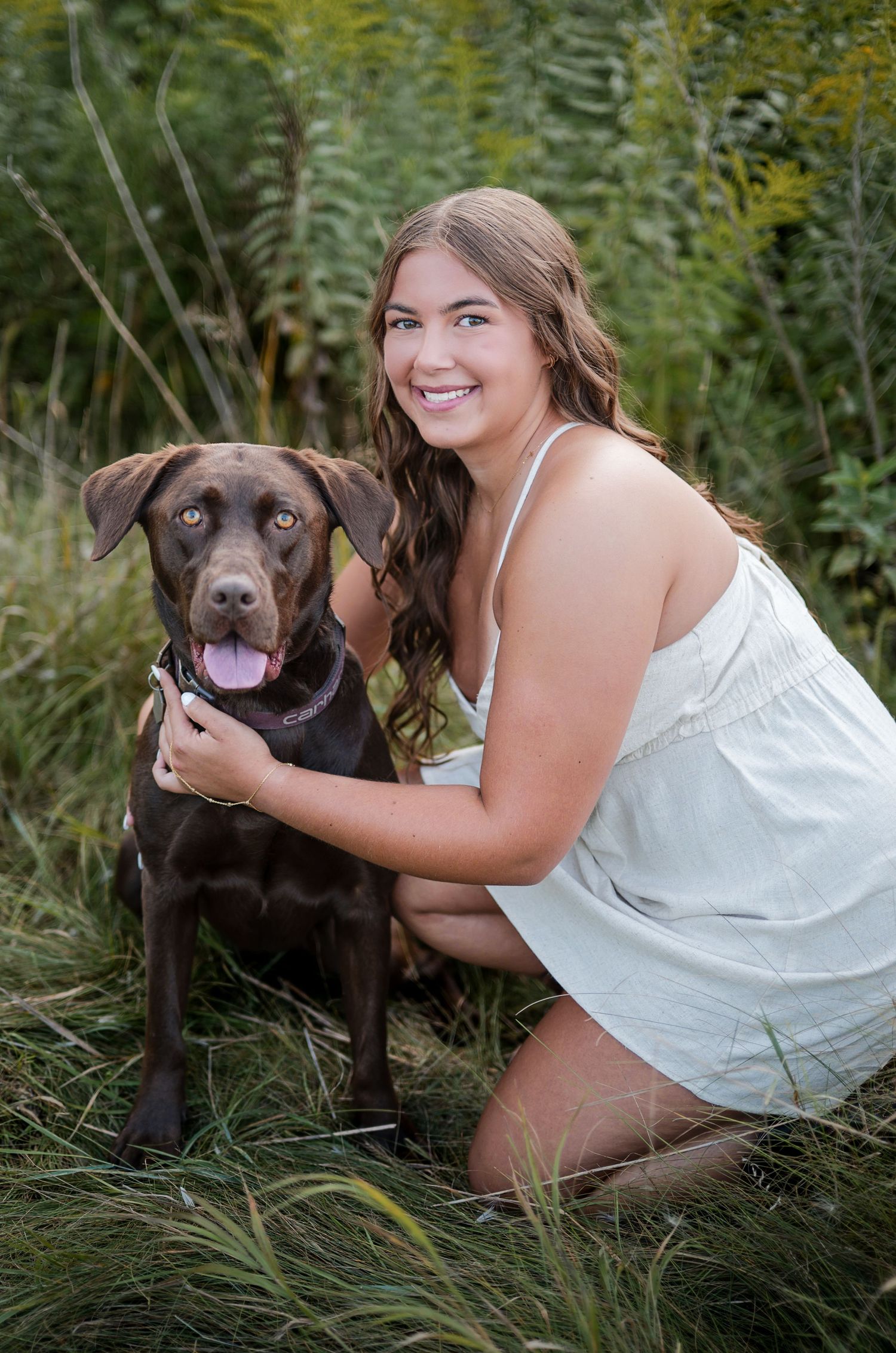 Young woman in white dress smiling with chocolate Labrador dog in lush green field outdoor portrait.