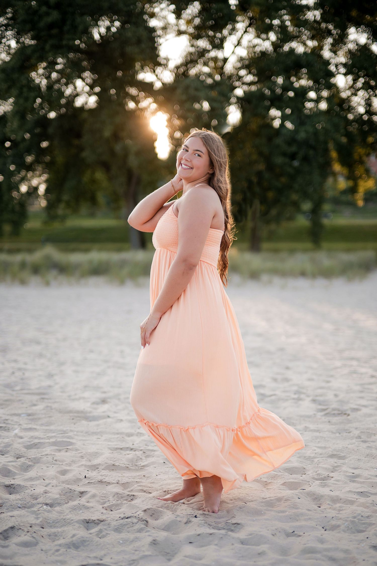 Woman in peach maxi dress twirling barefoot on sandy beach at golden sunset with trees in background.