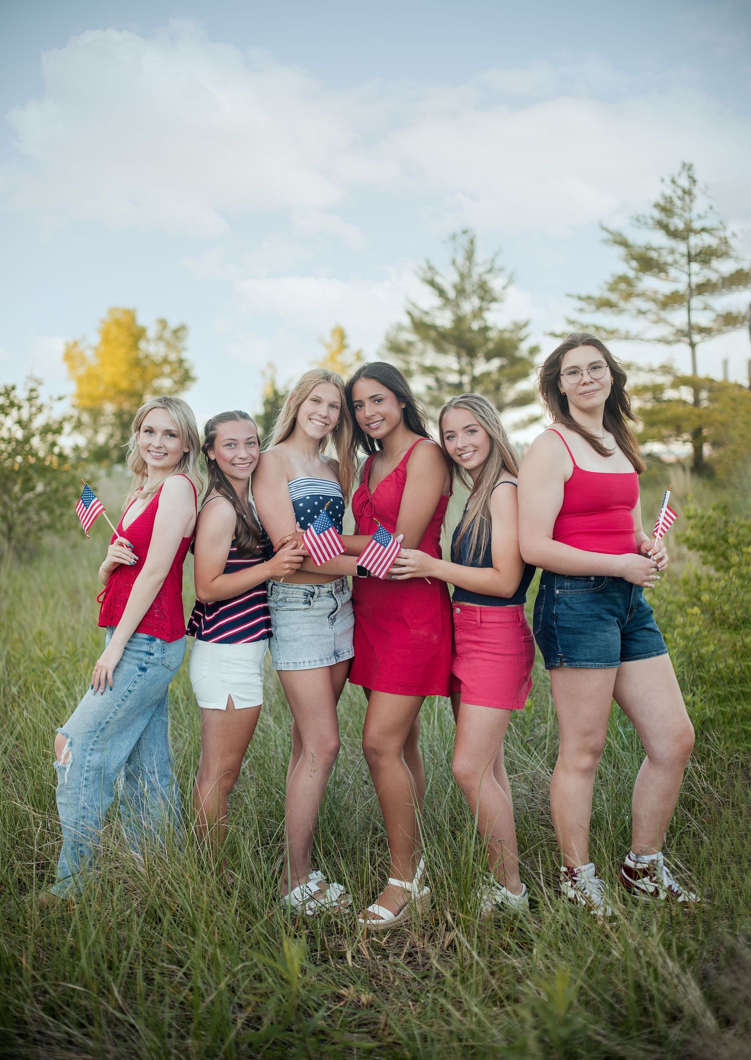 Six smiling girls in red, white and blue outfits hold small American flags while posing in a tall grassy field.